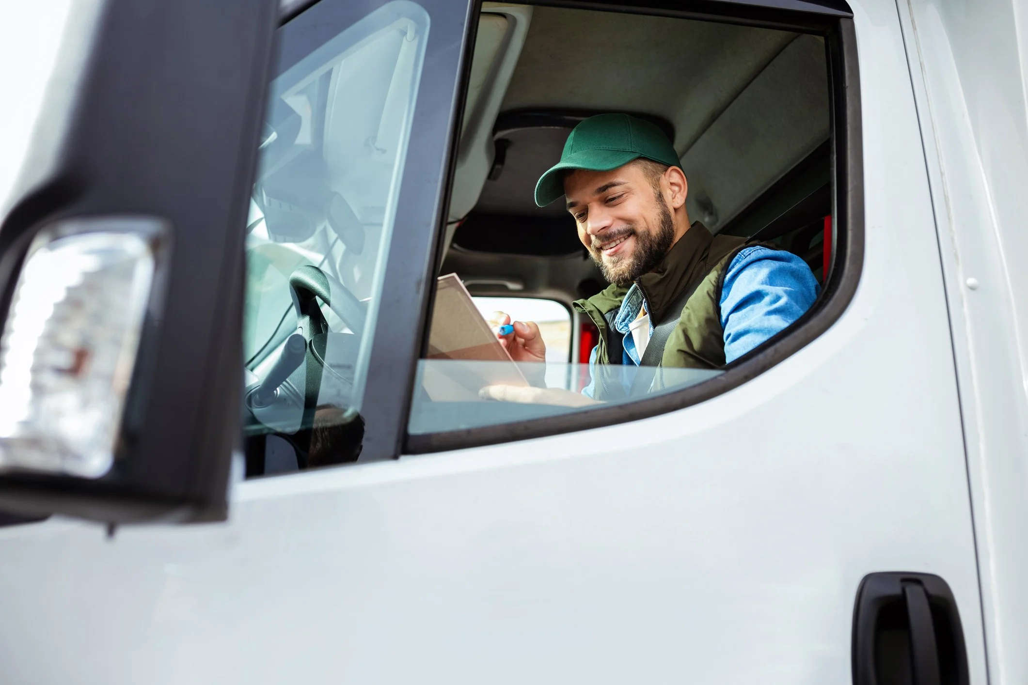 Smiling truck driver in a green cap and vest writes on a clipboard inside the vehicle, viewed through the window.