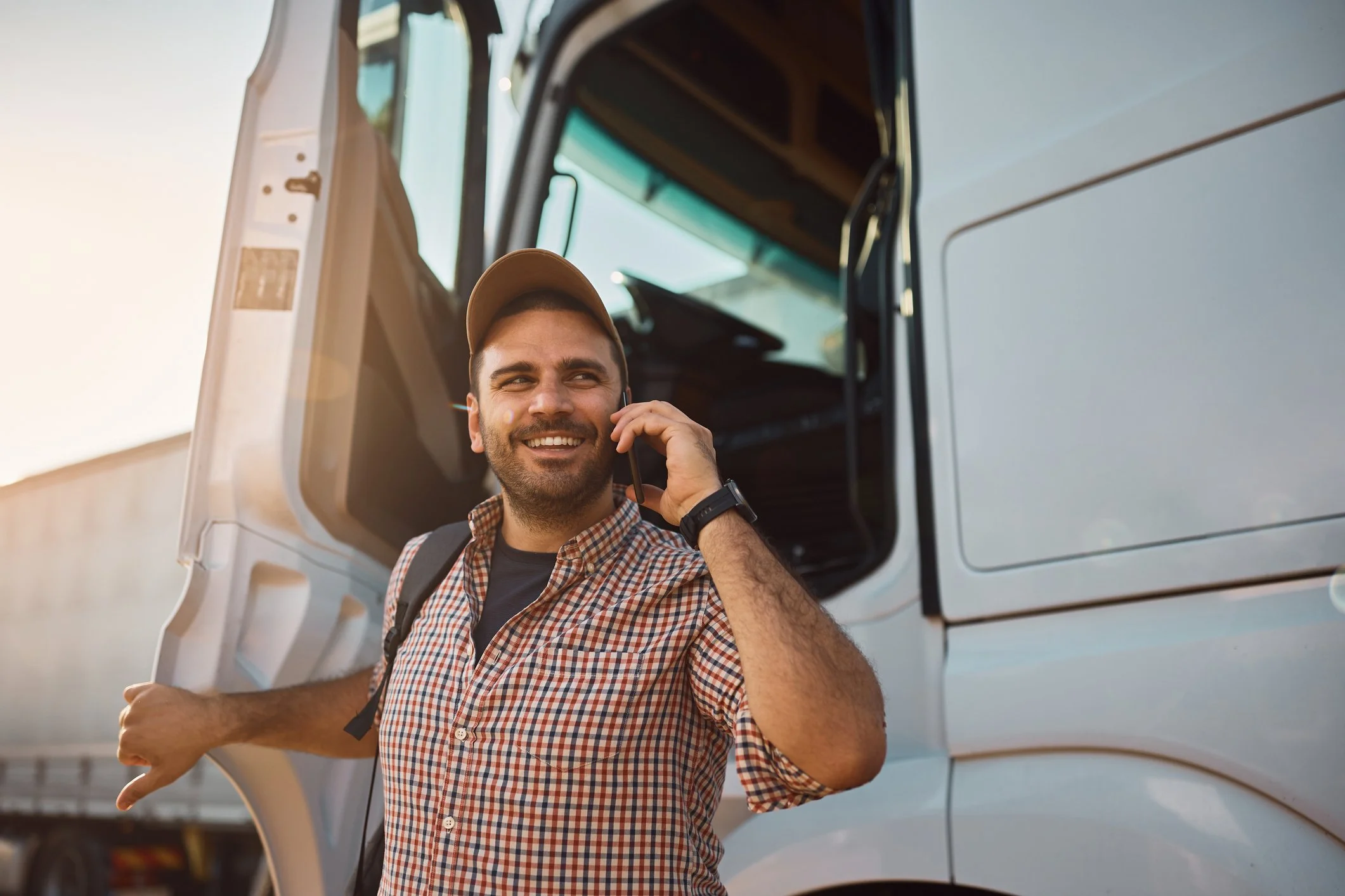 A man in a plaid shirt and cap stands at a truck's open door, smiling and talking on a phone.