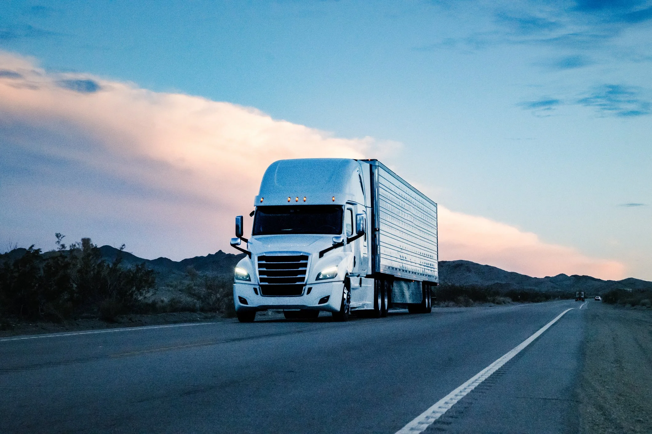White semi-truck on a desert highway at sunset, with a pink and blue sky.