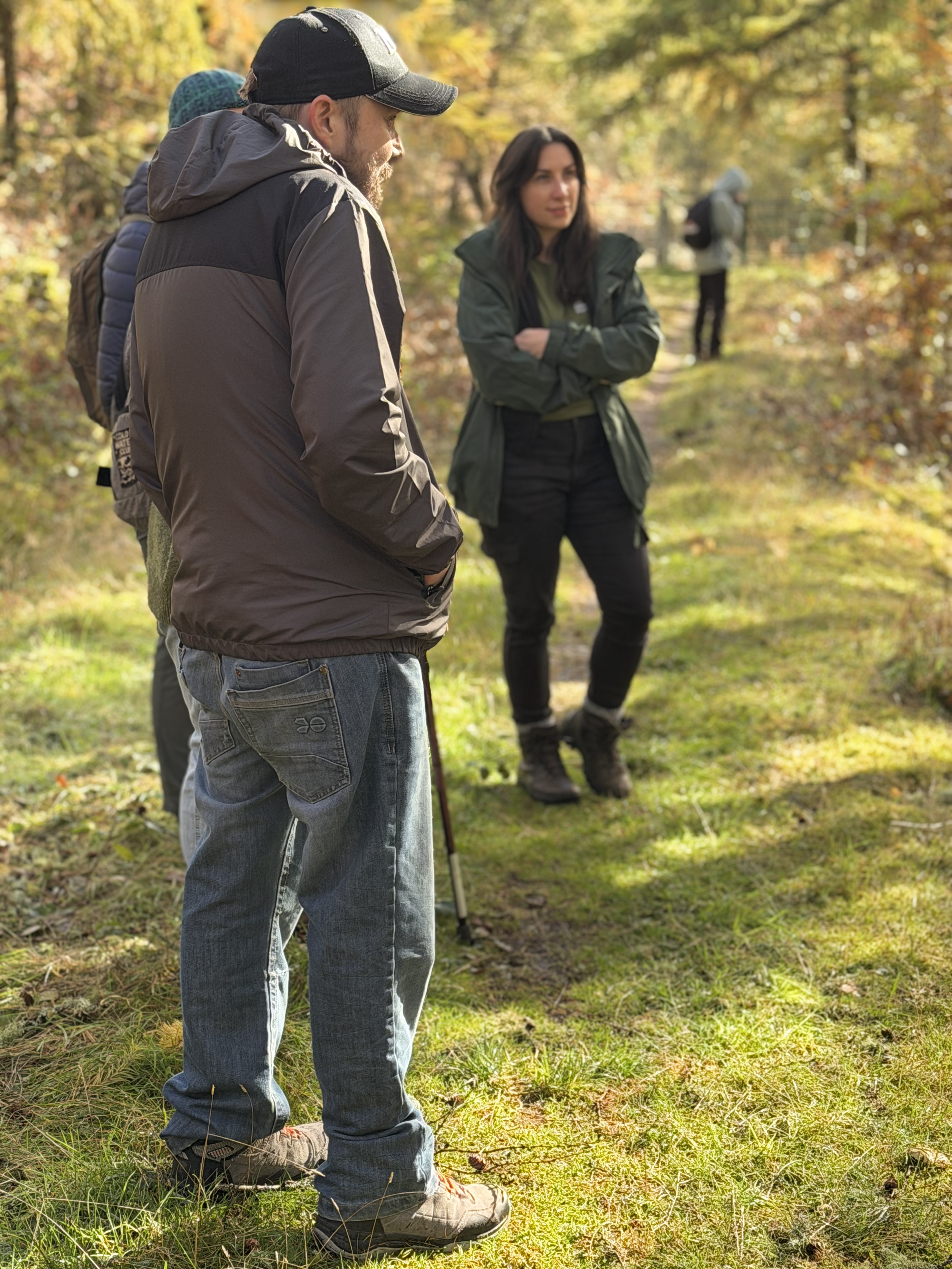 A group of people, including a man in a baseball cap and outdoor jacket, and a woman in a green jacket, standing on a forest trail during fall with colorful trees.