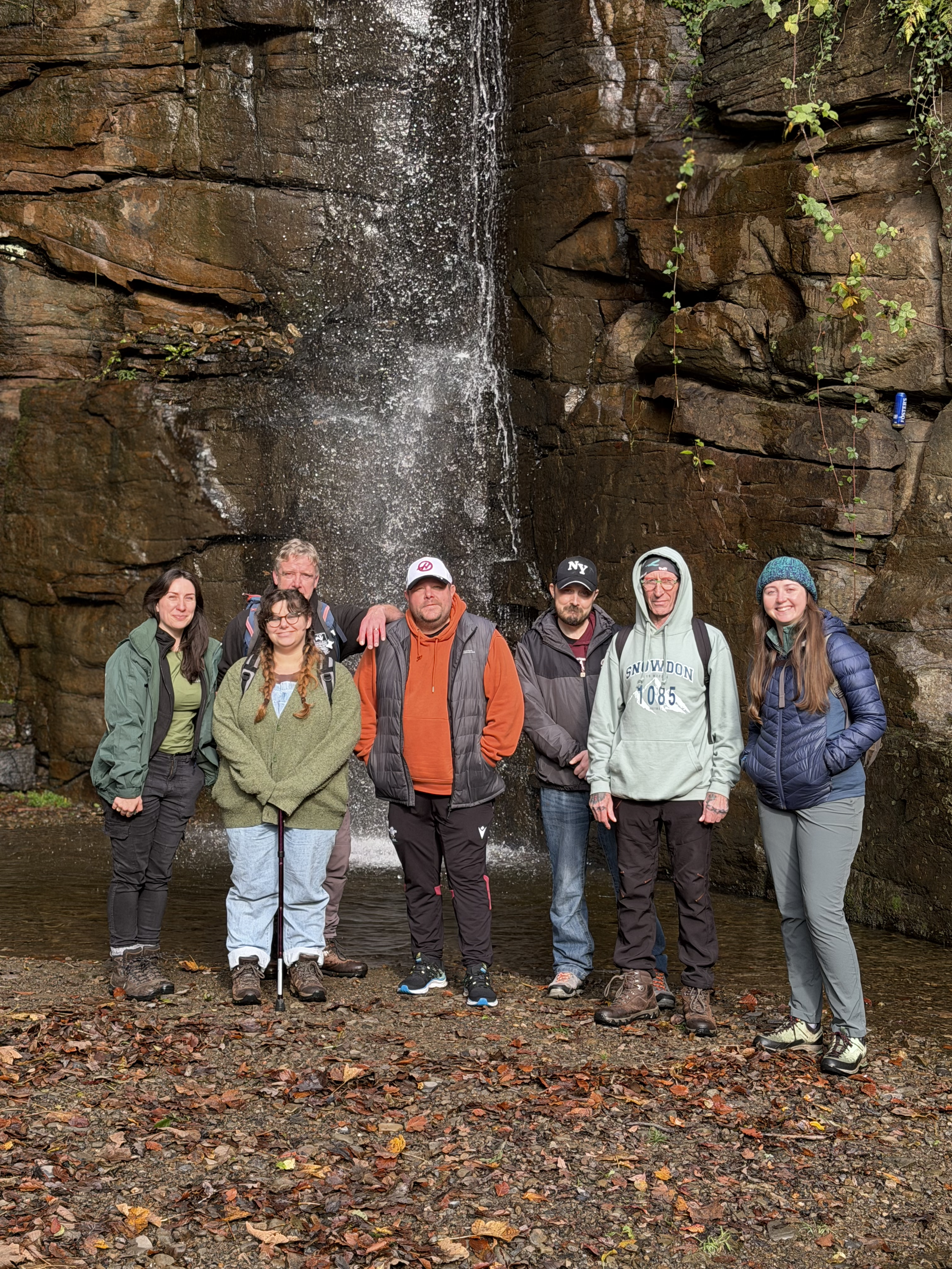 Group of seven people standing in front of a waterfall during a hike, with autumn leaves on the ground.