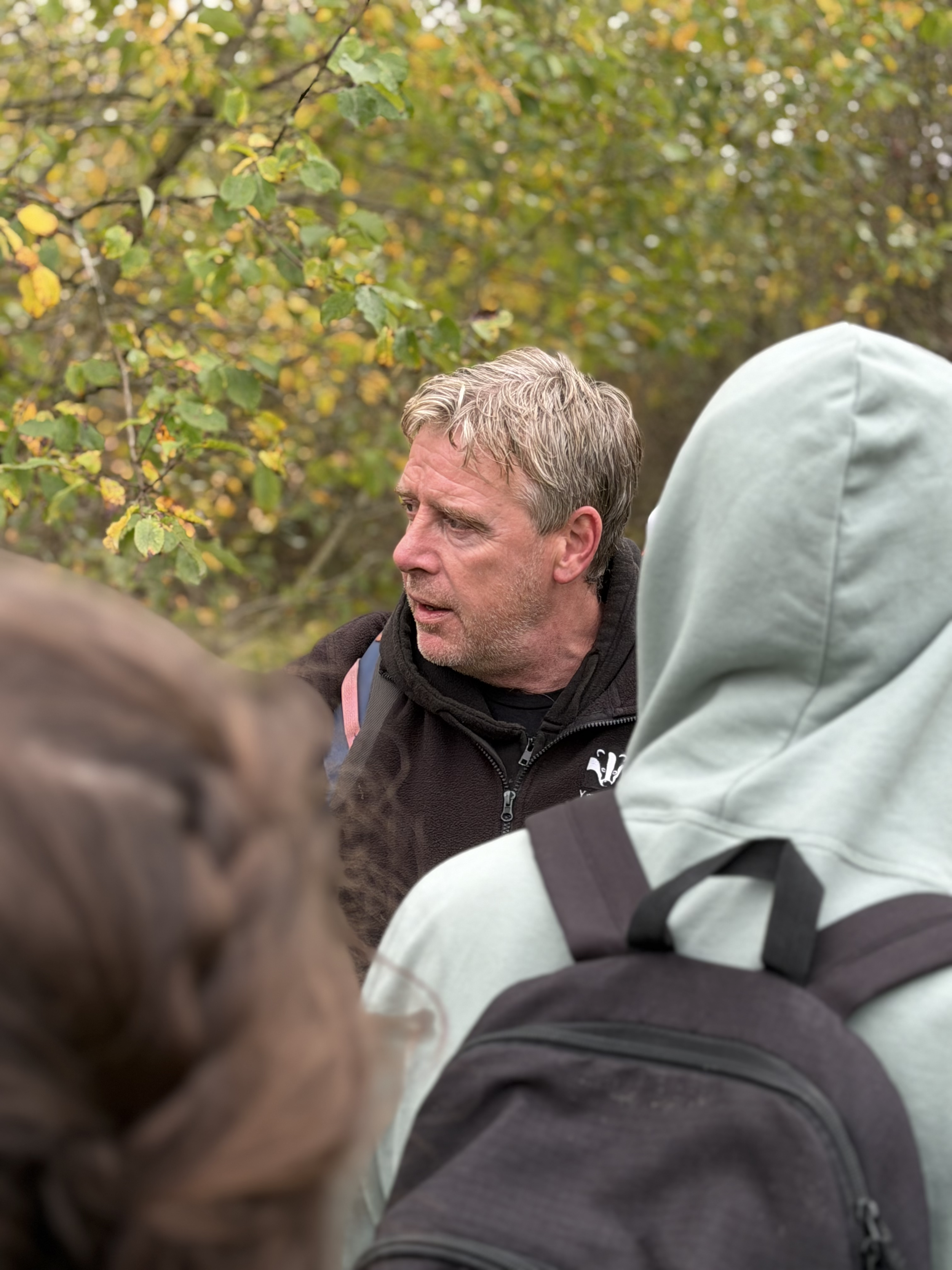 A man with blonde hair and a beard, wearing a black jacket, speaking outdoors to a group of people, with trees showing fall-colored leaves in the background. Part of a person in a gray hoodie and black backpack and a brown dog are also visible.