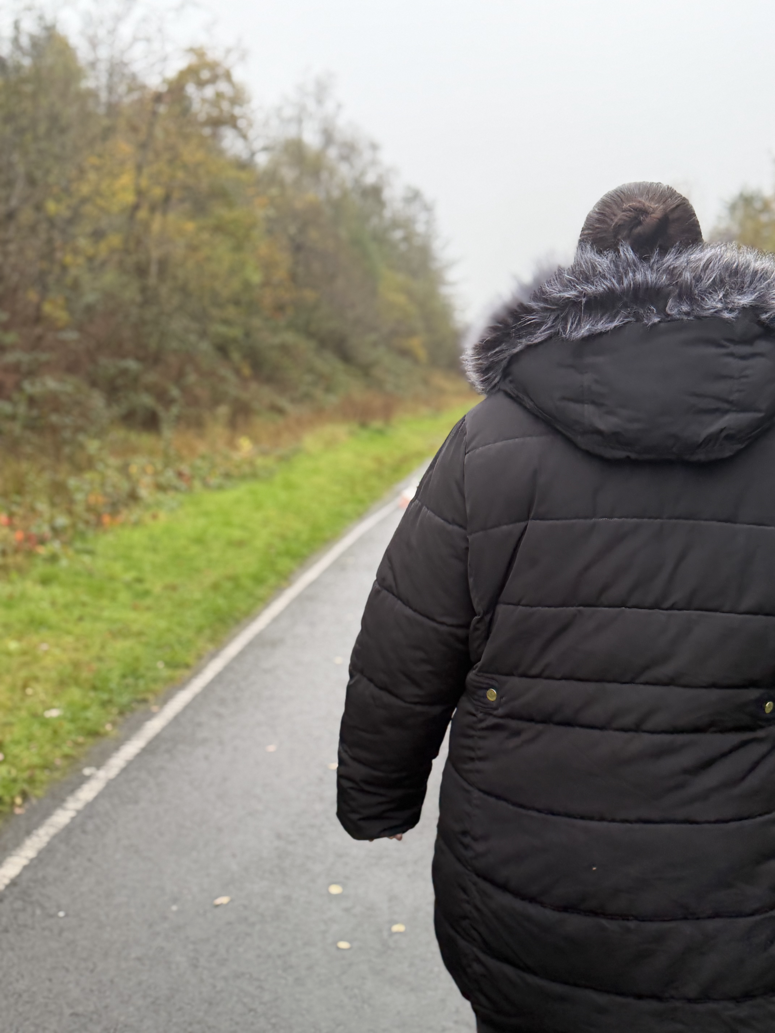 Person wearing a black winter coat with a fur-lined hood walking on a wet road surrounded by autumn trees.