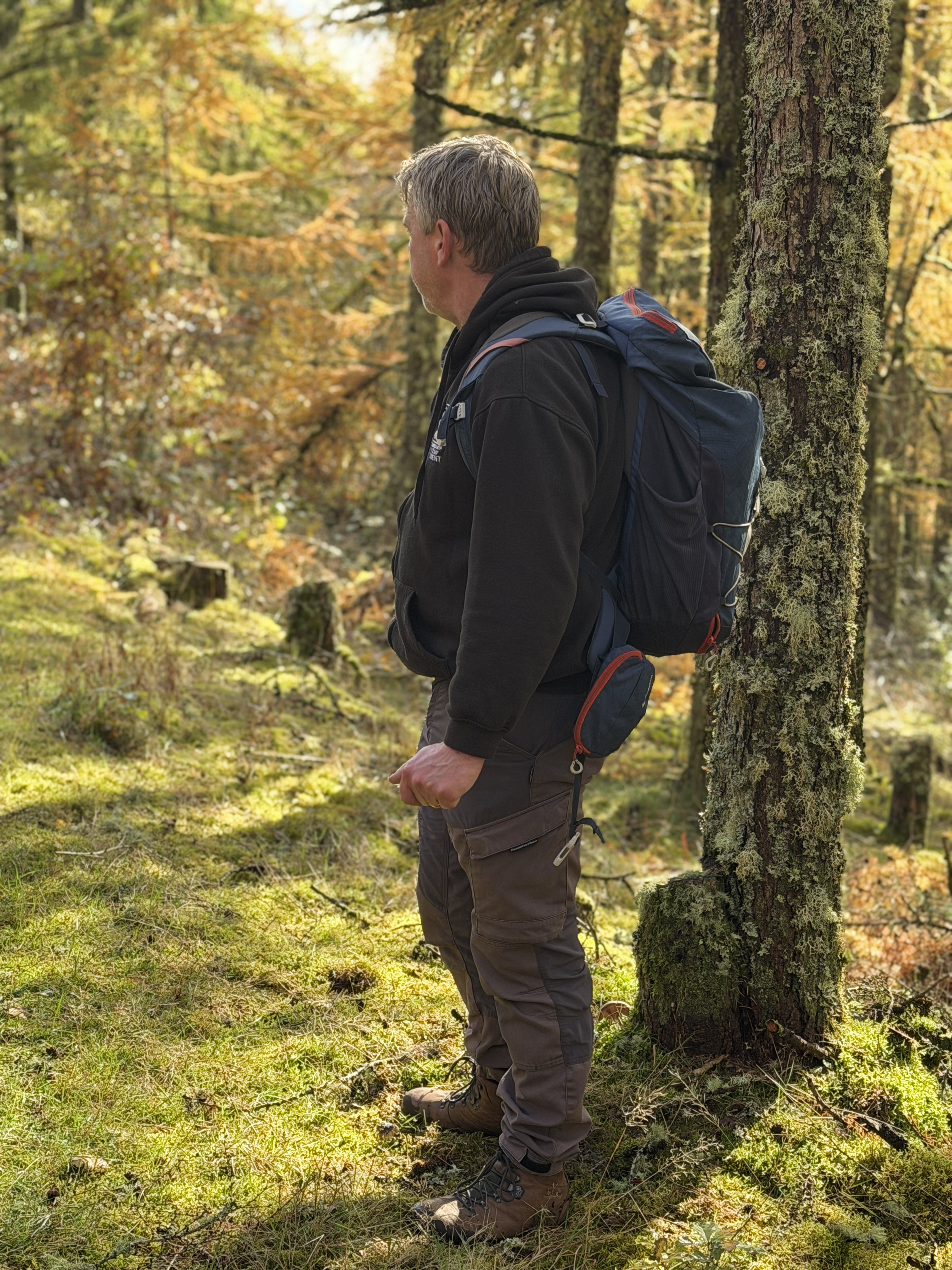 Man with gray hair, wearing black hoodie and brown hiking pants, standing in a forest with autumn-colored leaves, leaning against a moss-covered tree with a large blue backpack.