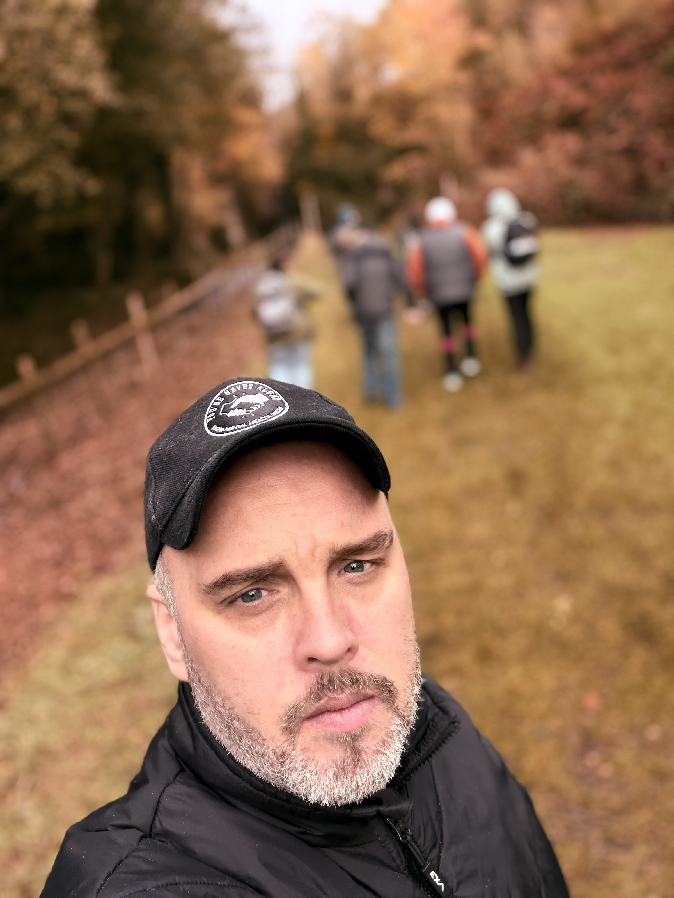 A man with a serious expression, wearing a black cap and jacket, taking a selfie outdoors on a fall day with a group of people walking in the background on a leaf-covered trail surrounded by colorful autumn trees.