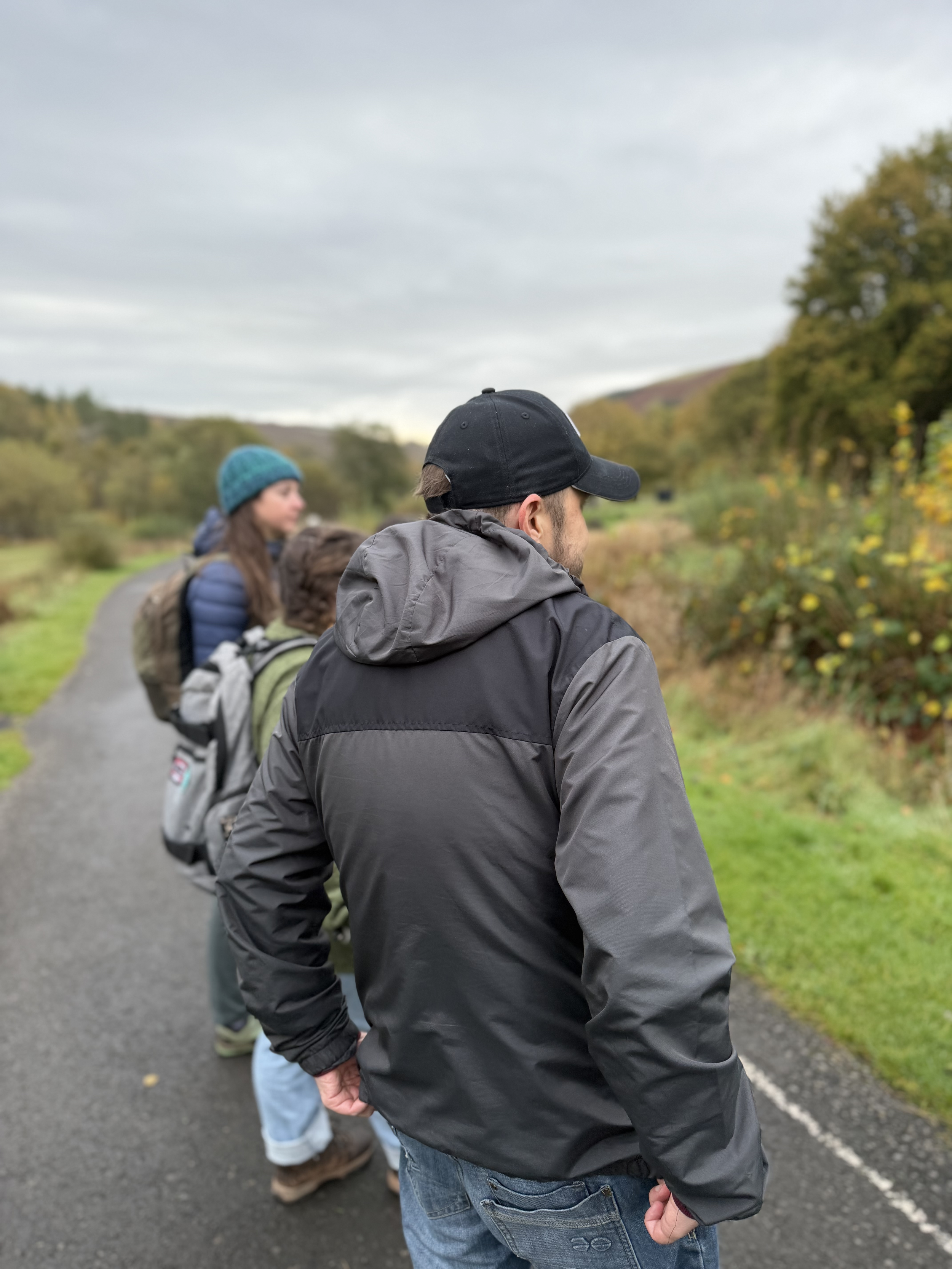 A group of people walking outdoors on a trail, with trees and shrubs on either side, under a cloudy sky.