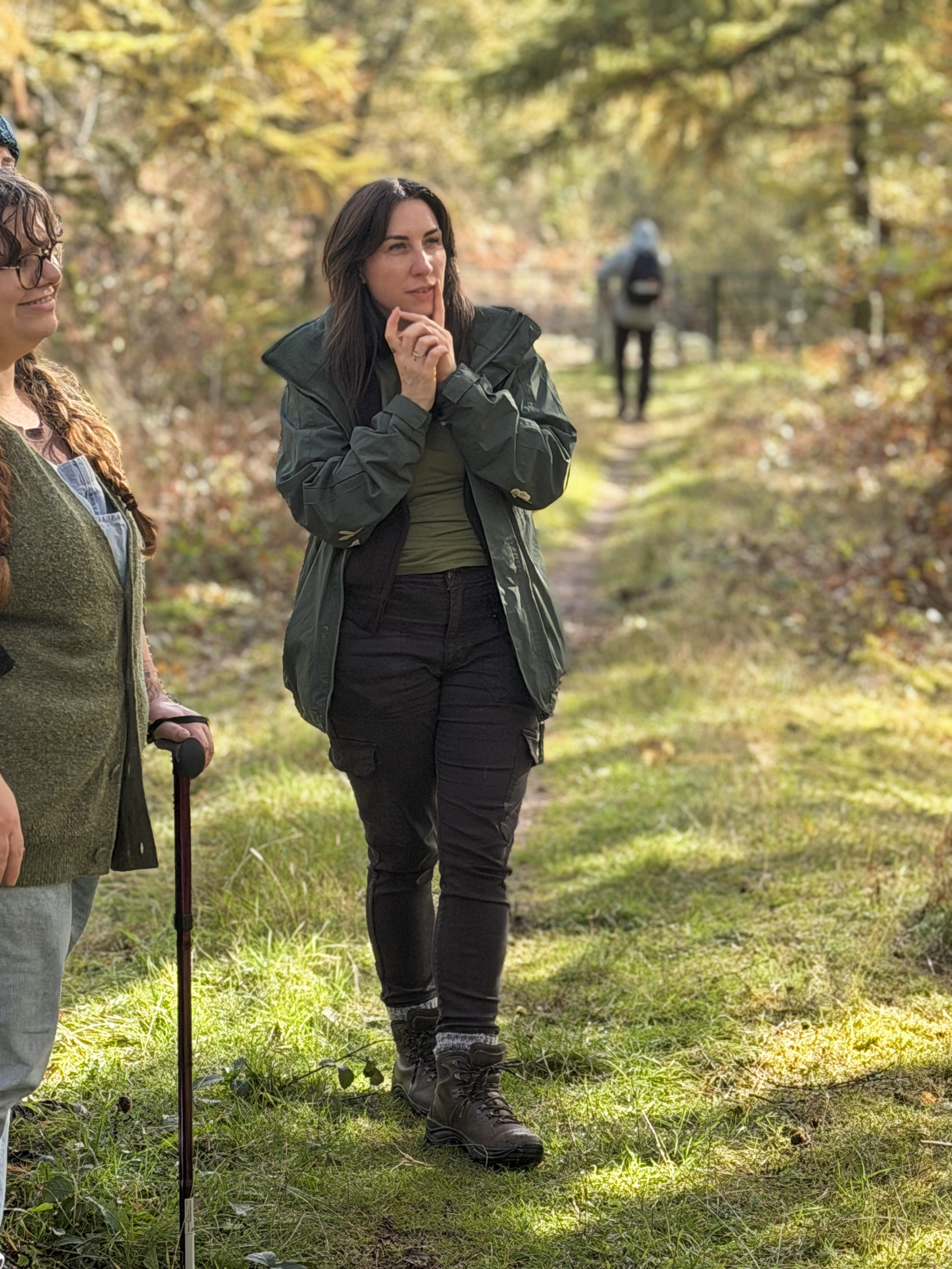 Women hiking outdoors on a trail in a forest during fall with colorful foliage, one woman in front wearing a green jacket and black pants, another woman partially visible with a walking stick, and a person with a backpack in the background.