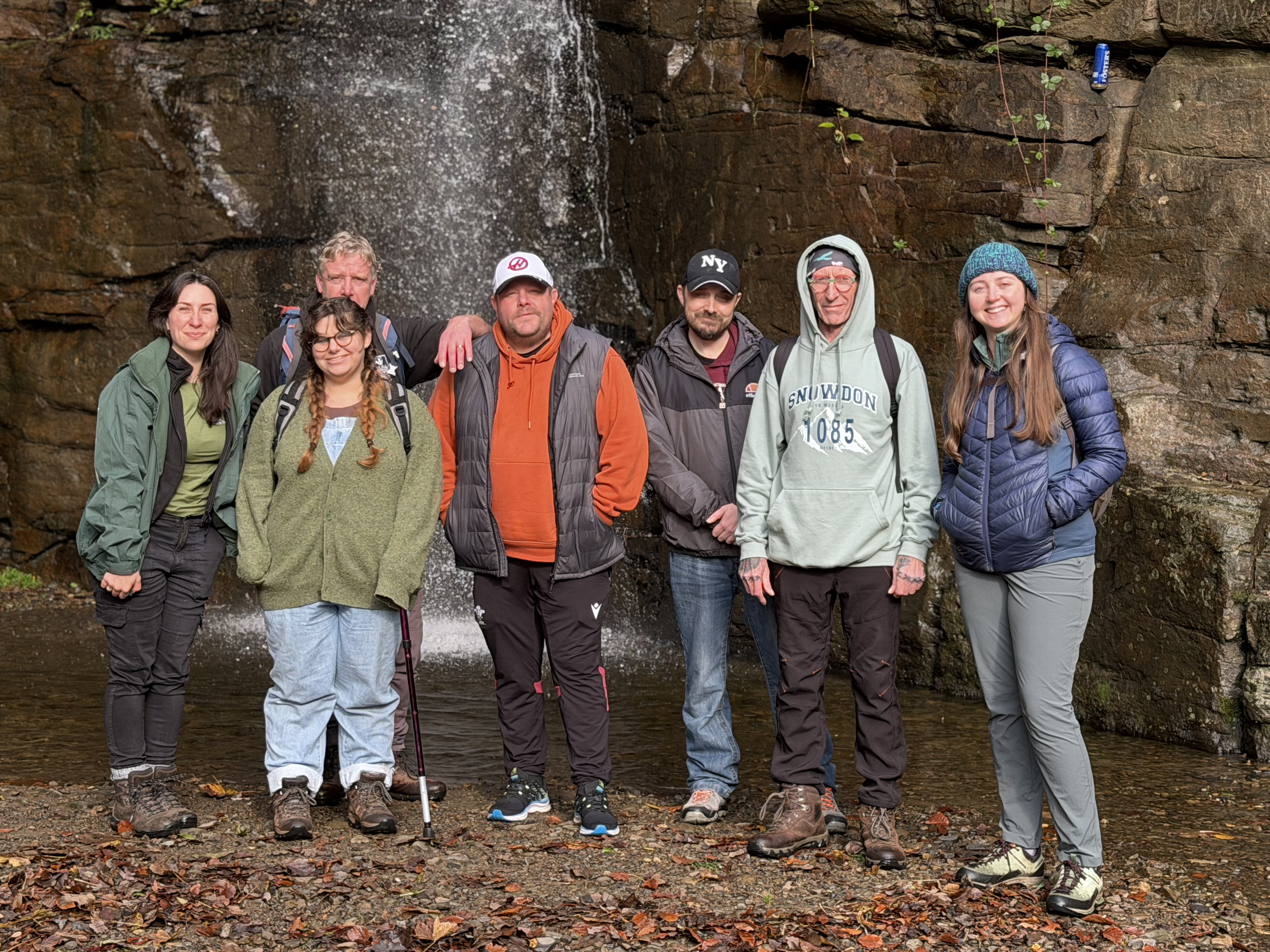 Group of seven people standing outdoors near a waterfall, dressed in hiking gear, smiling at the camera.