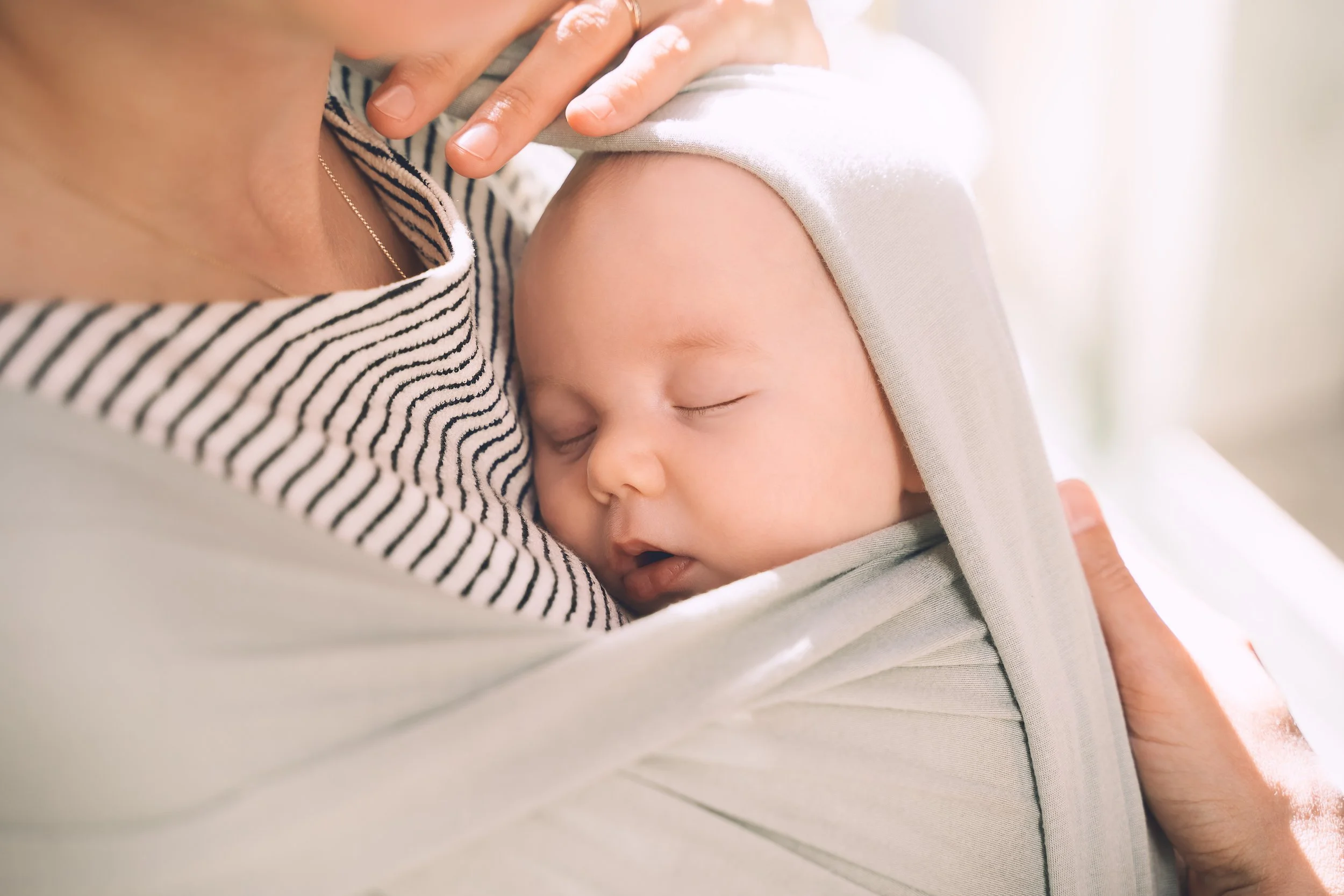A baby sleeping peacefully against a woman's chest, with the woman gently holding the baby's head. The baby is wearing a beige hat and pajamas, and the woman is wearing a striped shirt.