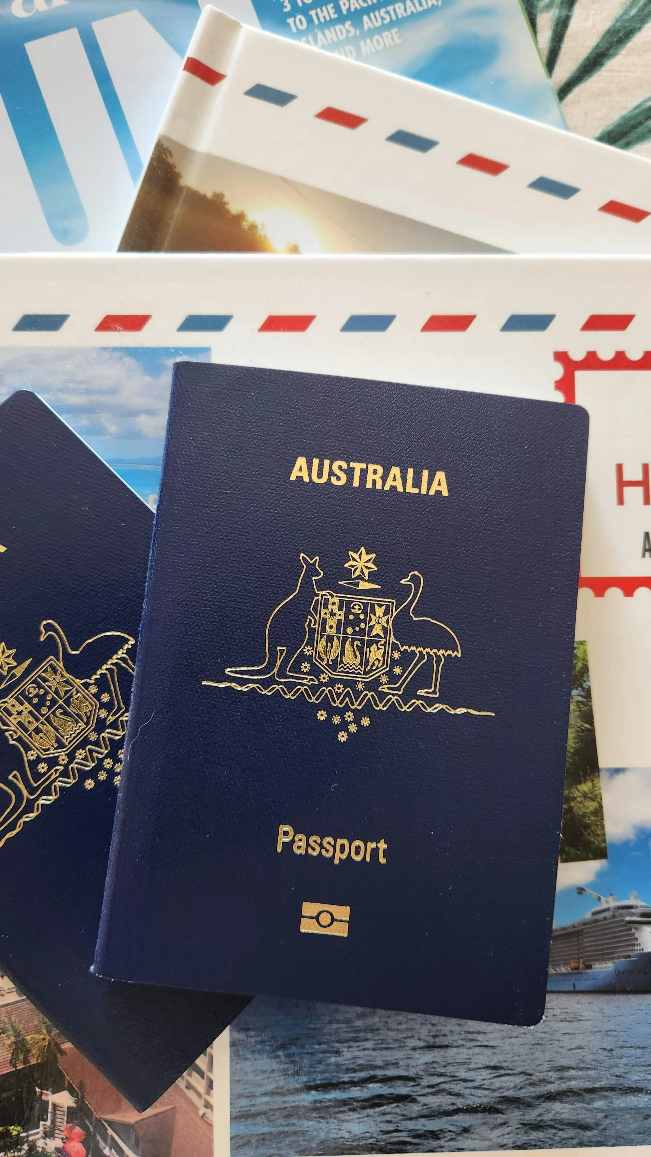 Australian passport resting on travel brochures and postcards. The brochure with a sunset and ocean scene, and postage stamp designs in red and blue.