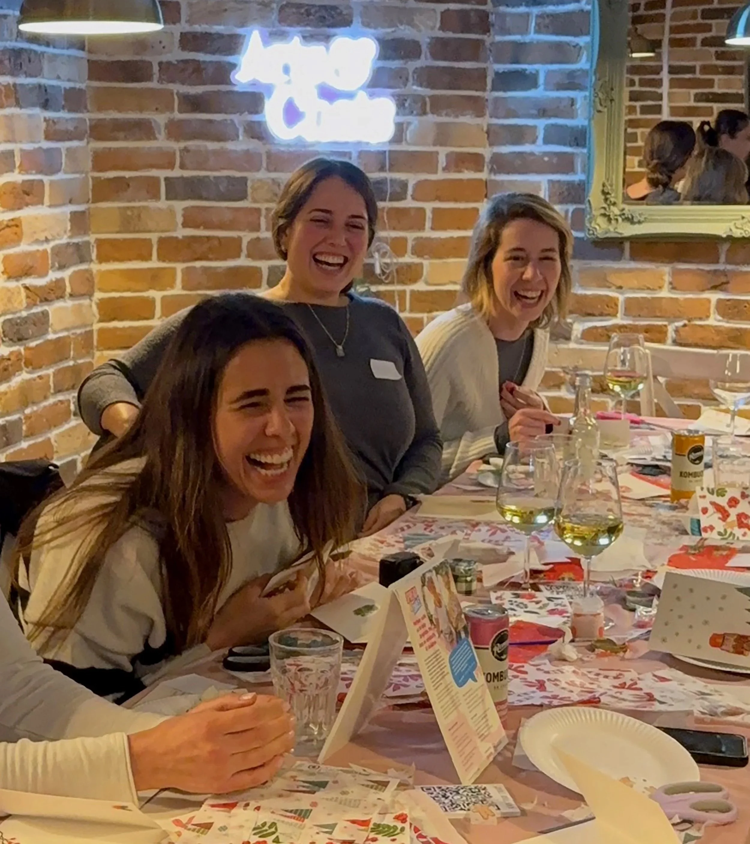 Three women sitting at a table laughing and having fun, with a brick wall and a neon sign in the background.