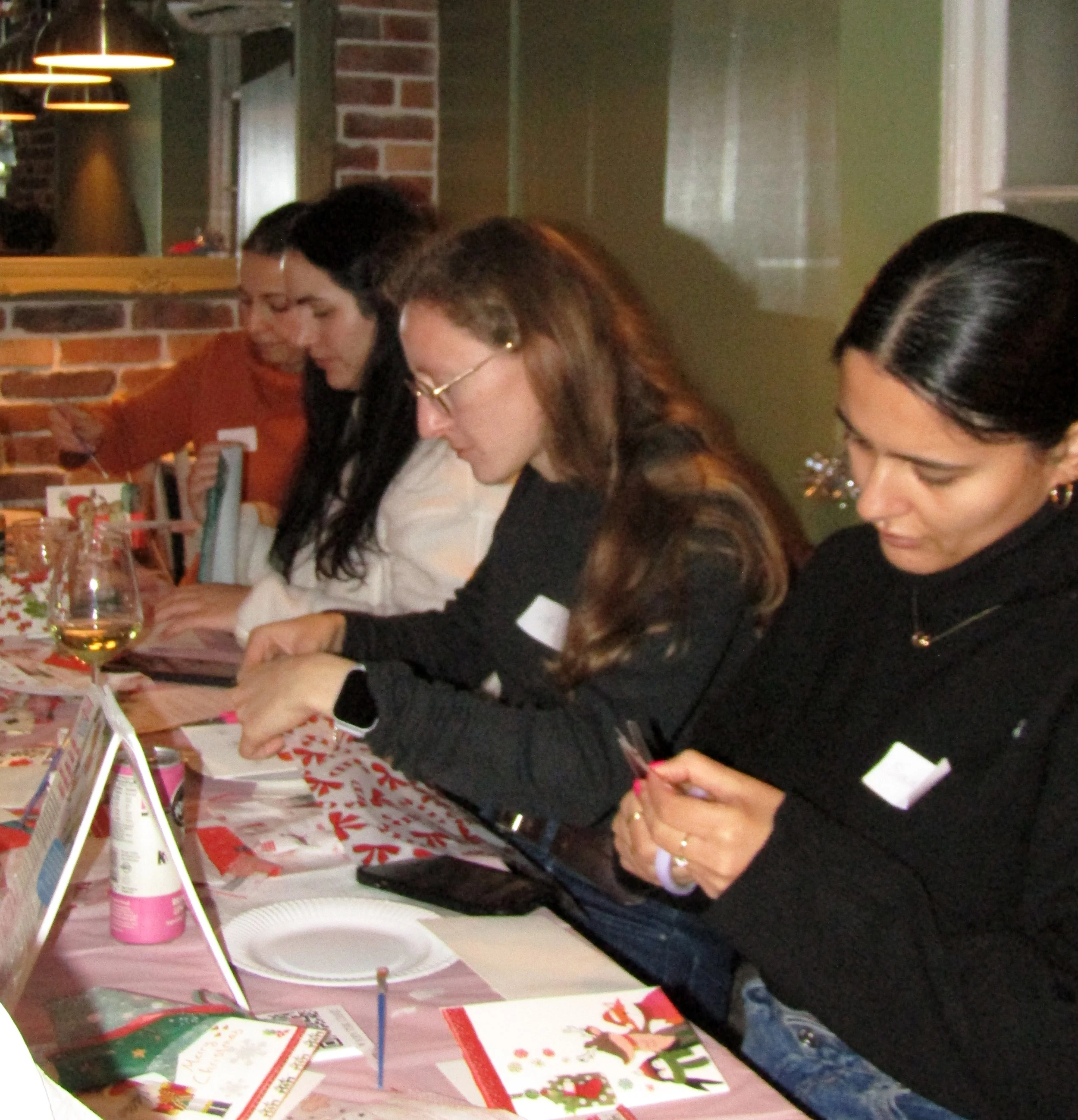 Four women sitting at a decorated table, engaged in holiday activities such as reading cards and writing, with Christmas-themed decorations and tableware.