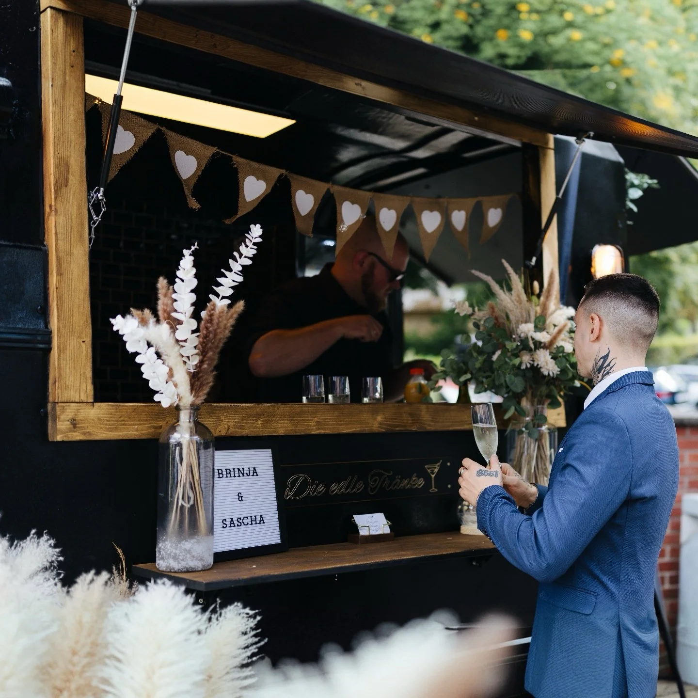 Ein Mann in einem blauen Anzug mit Tattoo am Hals hält ein Glas Sekt und bestellt an einem mobilen Getränkestand mit Holzkonstruktion und dekorativen Blumen. Im Verkaufswagen steht eine Person, die Getränke zubereitet.