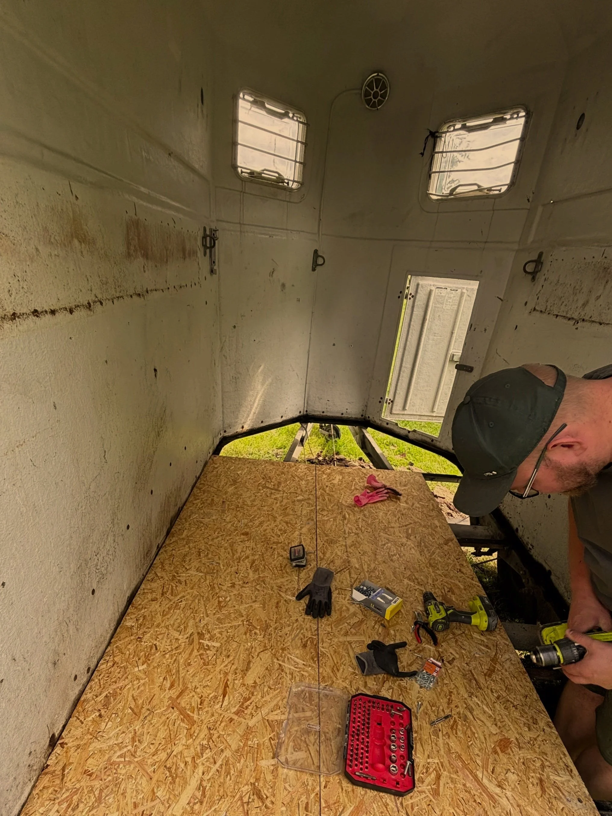 Innenansicht einer kleinen, ungenutzten Segelyacht mit offener Tür und zwei kleinen Fenster, Werkzeuge und Ausrüstung auf einer Holzplatte, eine Person mit schwarzer Kappe hält ein Akkuschrauber.