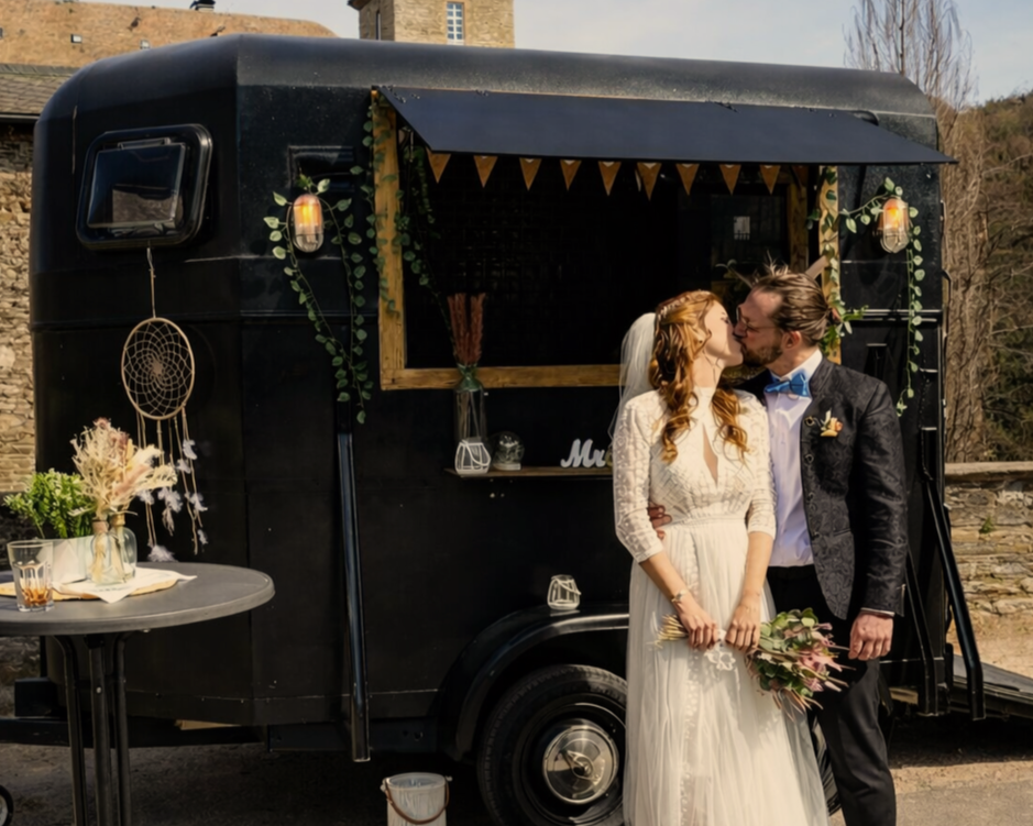 Ein Brautpaar küsst sich vor einem schwarzen Foodtruck bei einer Hochzeit, dekoriert mit Pflanzen und Lichterketten.