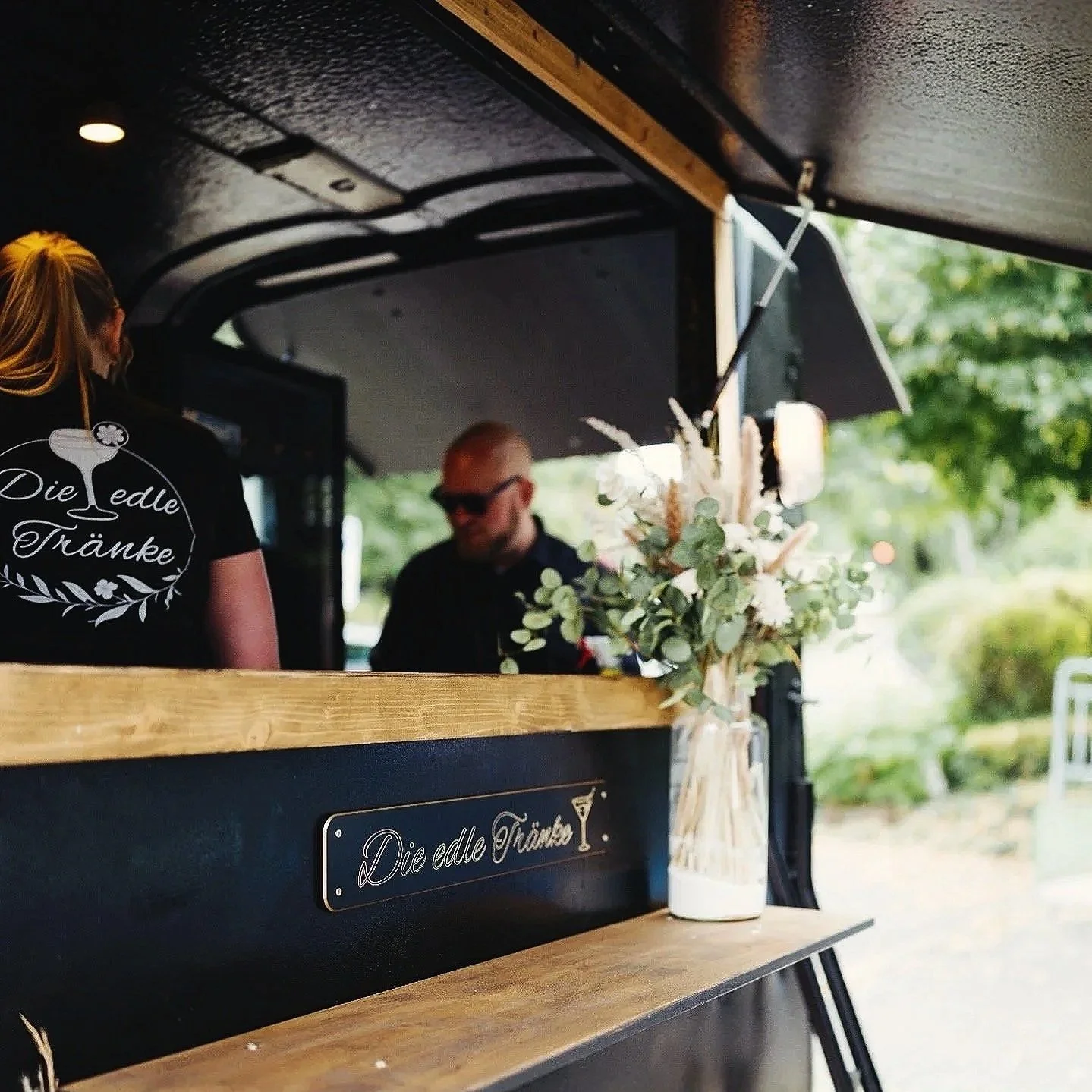 Blick auf einen mobilen Barwagen mit einem dekorativen Blumenstrauß in einer Vase, im Hintergrund eine Frau mit schwarzen T-Shirt mit goldener Schrift und einem Mann mit Sonnenbrille, im Freien bei Tageslicht.