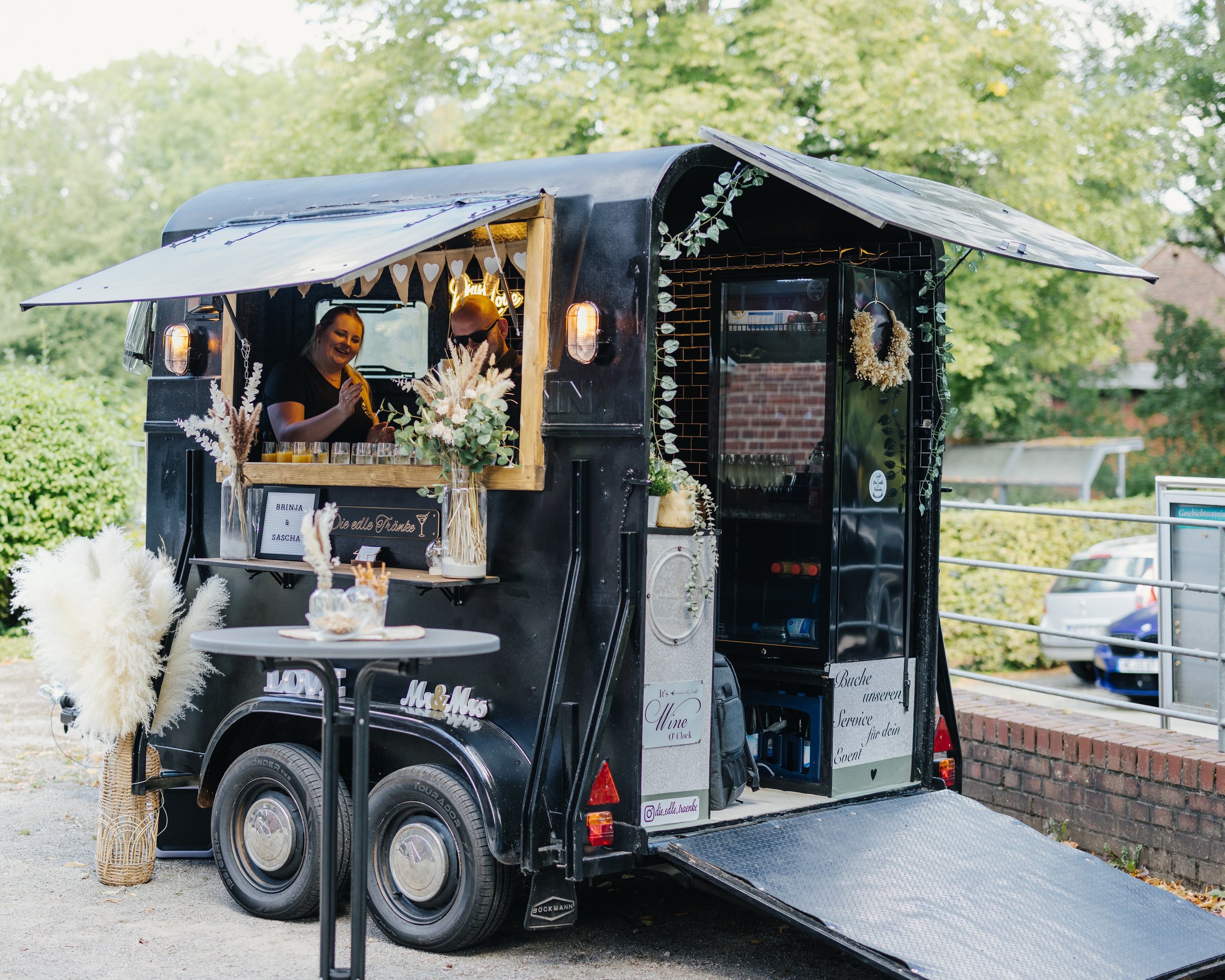 Ein schwarzer Food-Truck mit dekorativen Pflanzen und Herzgirlanden im Freien bei Sonnenschein, mit zwei Menschen, die Getränke und Snacks servieren.