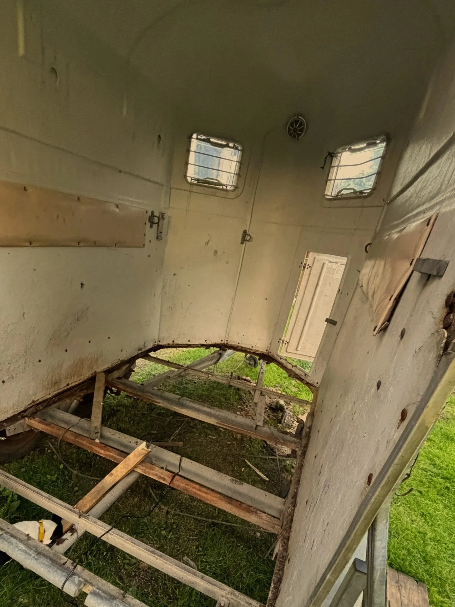 Innenansicht eines verfallenen Wohnwagens mit Moos und Schmutz auf den Wänden, einem geöffneten Fenster, einem herausgenommenen Bettgestell und einem beschädigten Boden.