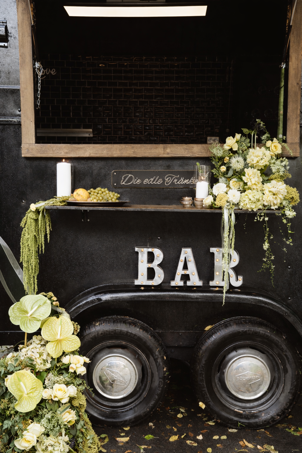Ein schwarzer Barwagen mit dekorativen Blumen, Kerzen, Obst und einem Lichtbuchstaben 'BAR' im Vintage-Stil. Es gibt eine Tafel mit der Aufschrift ‚Die edle Tränke‘, umgeben von Pflanzen und Dekorationen.
