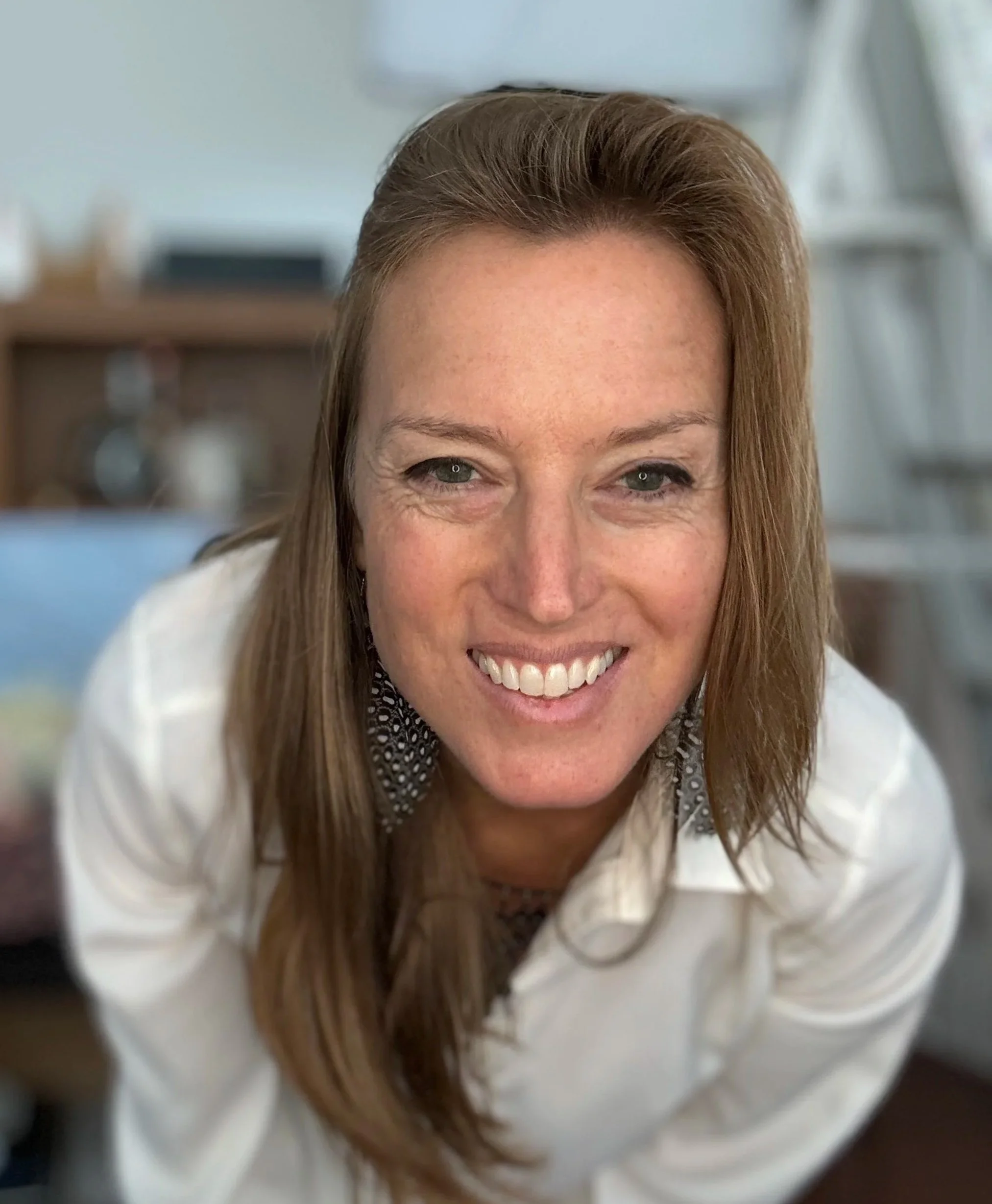 A woman with light skin, reddish-brown hair, and a white shirt smiling at the camera in a well-lit room.