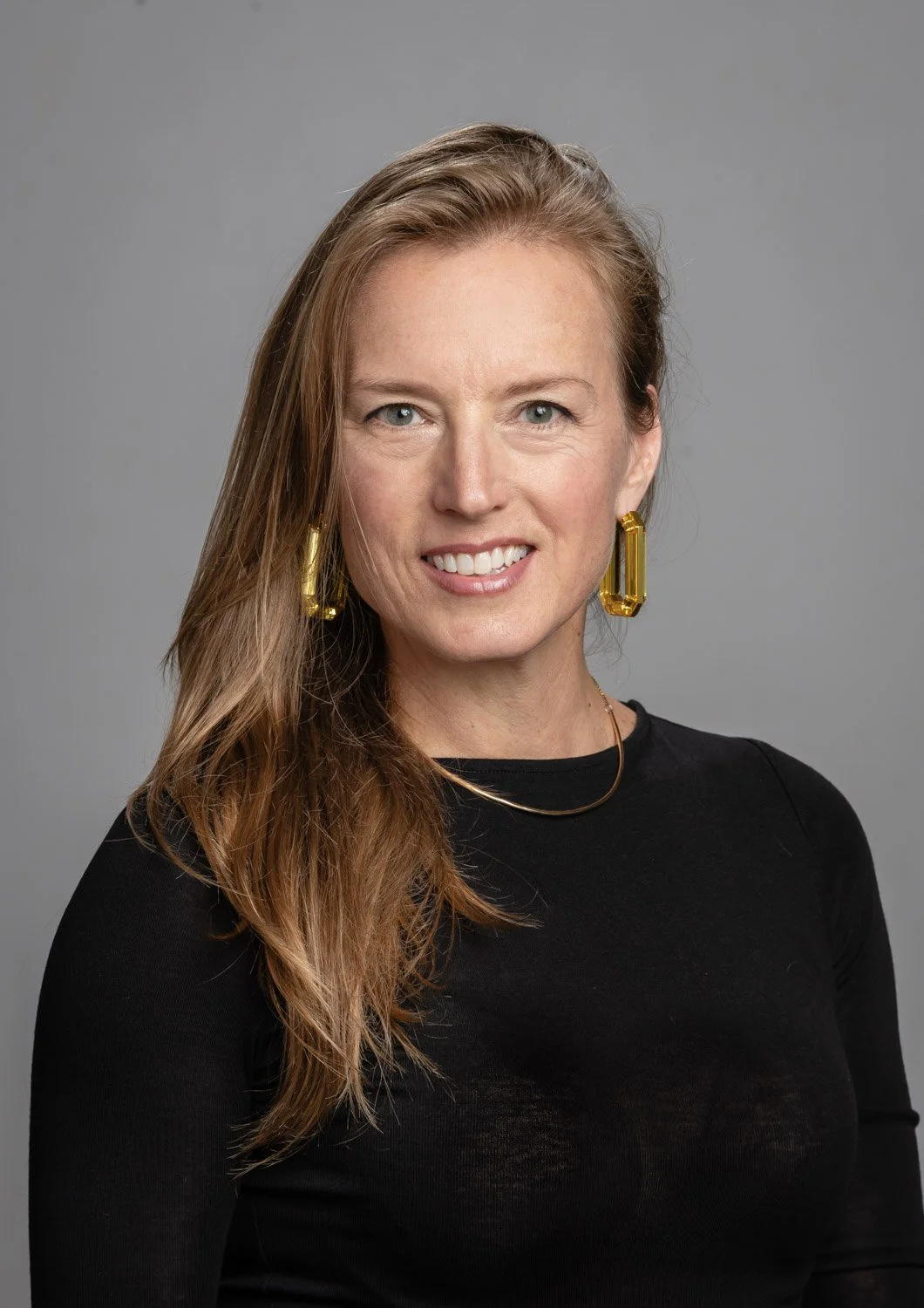 A woman with long red hair, wearing a black top, gold earrings, and a gold necklace, smiling at the camera against a gray background.