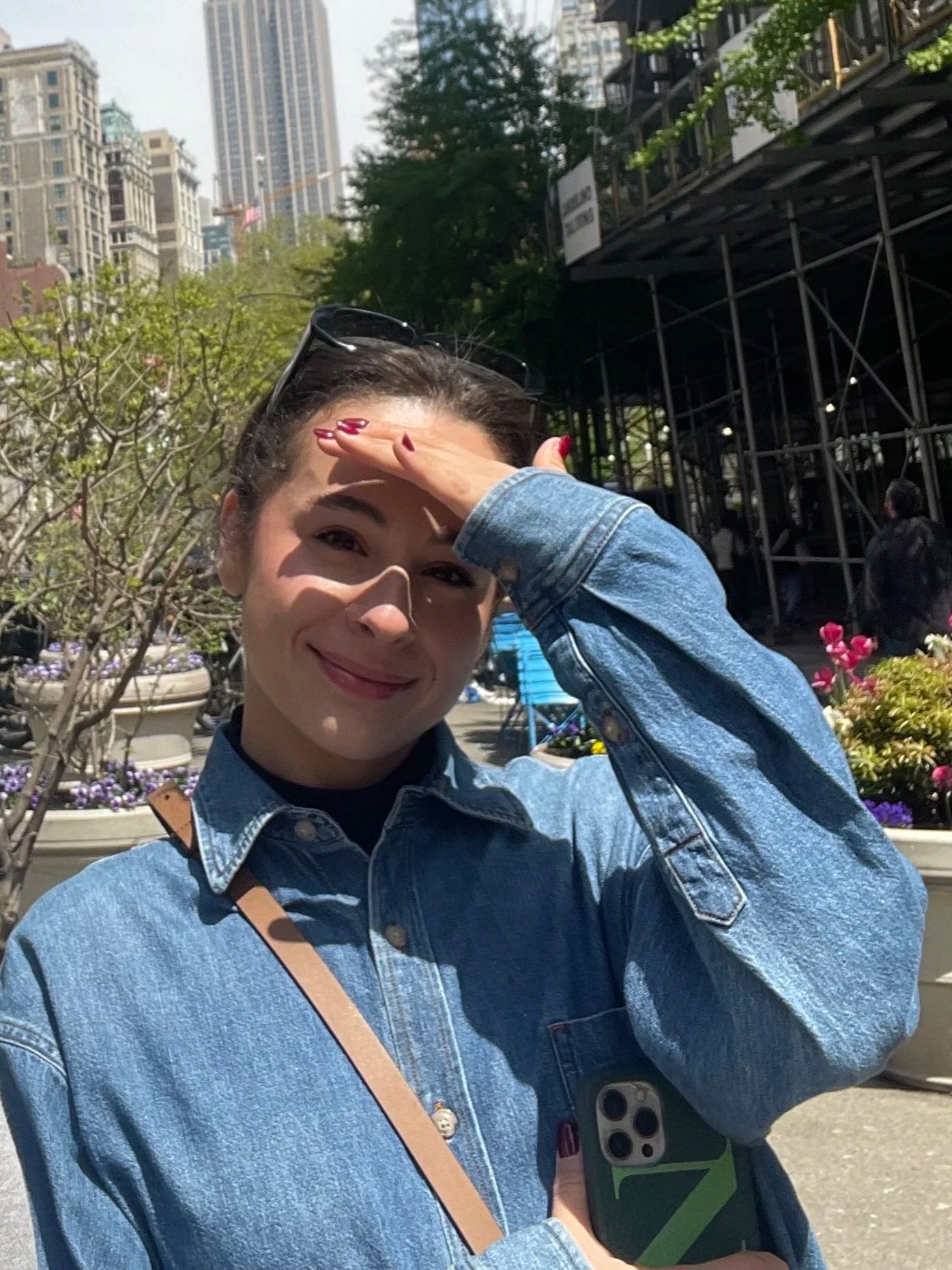 A young woman with dark hair, smiling, wearing a denim jacket and sunglasses on her head, shielding her eyes from the sun with her hand, standing outdoors in an urban park with flowers, trees, and tall buildings in the background.