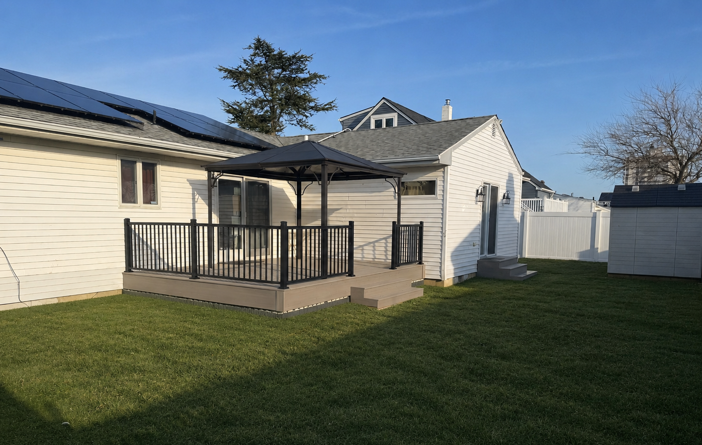 A backyard with a small wooden deck featuring black railing and a gazebo, surrounded by green grass and white fences, with neighboring houses in the background under a clear blue sky.
