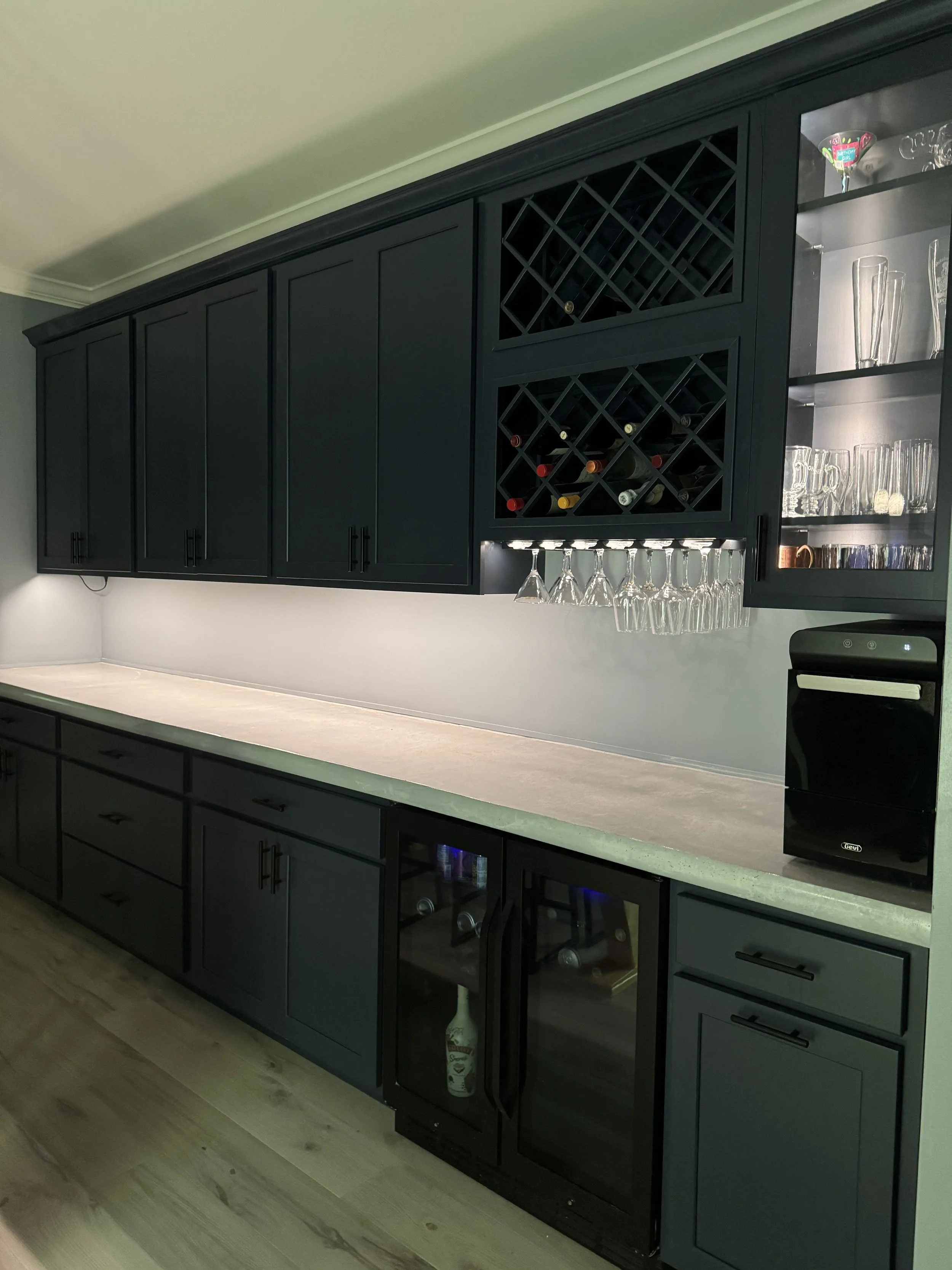 Black kitchen cabinets with a light-colored countertop, wine rack, glass storage, and a mini wine fridge, in a modern kitchen.