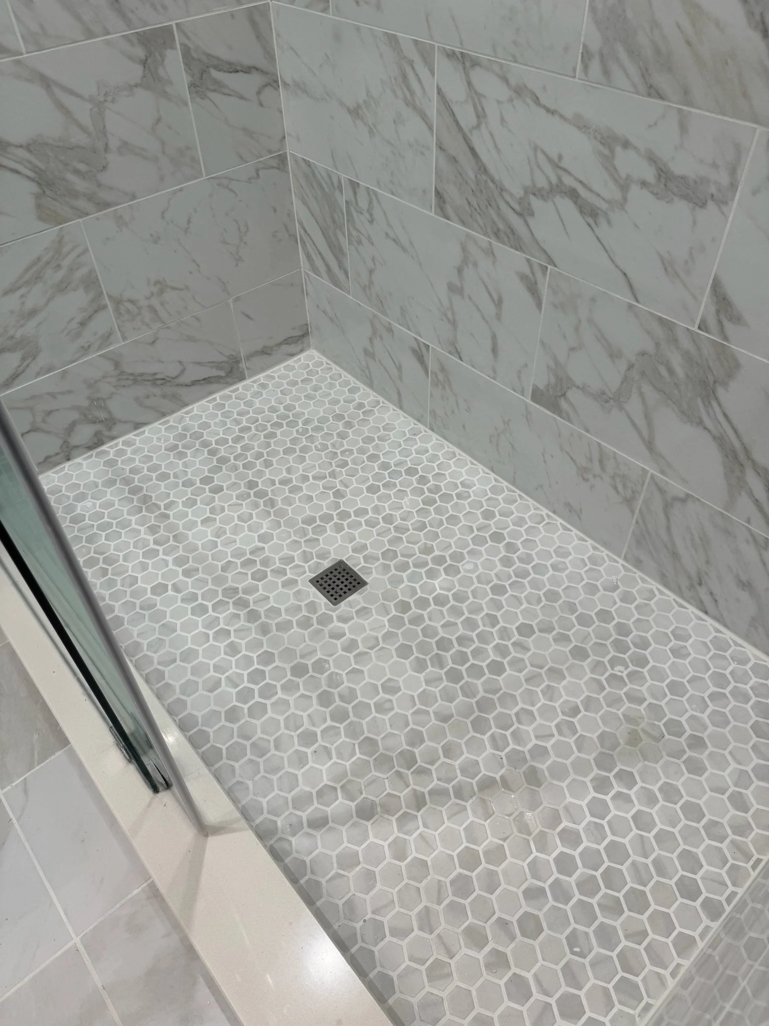 Shower area with large marble-patterned tiles on the walls and small white hexagonal tiles on the floor, including a drain in the corner.