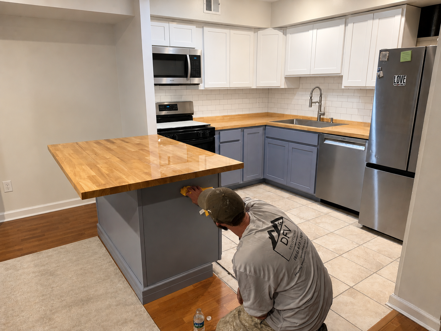 A person working under a granite kitchen island in a modern kitchen, with white upper cabinets, blue lower cabinets, stainless steel appliances, and a wooden countertop.