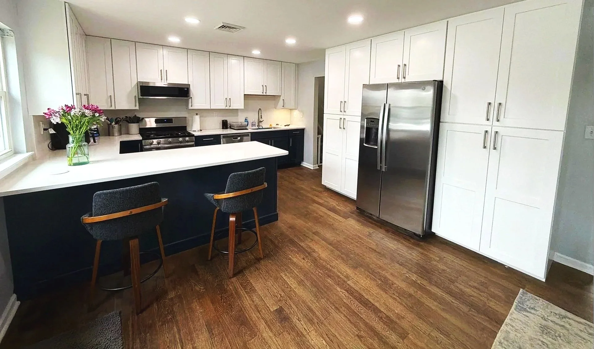 Modern kitchen with white cabinets, stainless steel refrigerator, dark island with two bar stools, and hardwood flooring, with a flower vase on the counter and a window on the left.