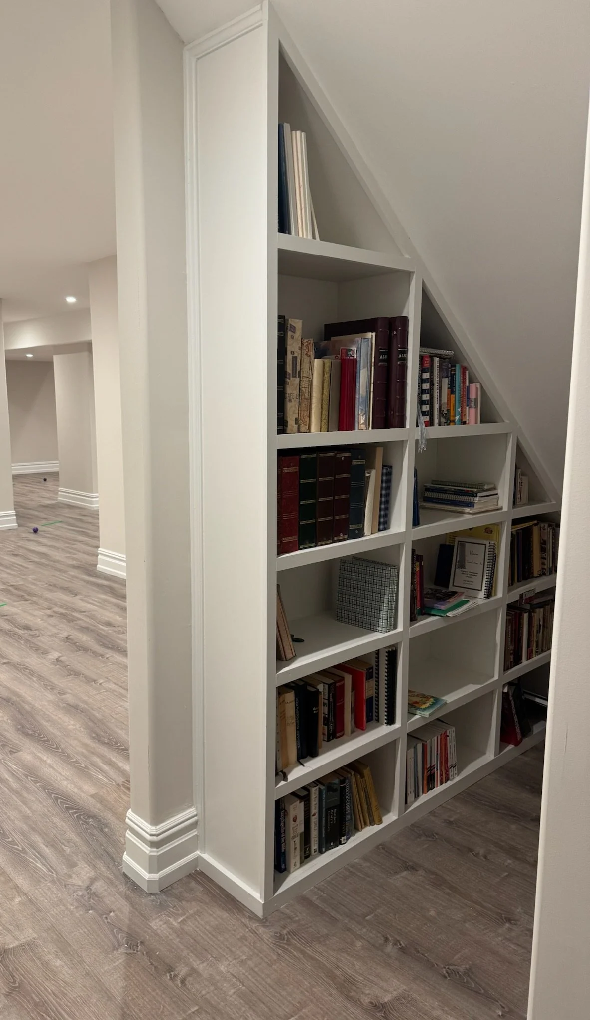 White bookshelf with multiple shelves filled with books, positioned against a white wall in a room with wood-effect flooring and sloped ceiling.