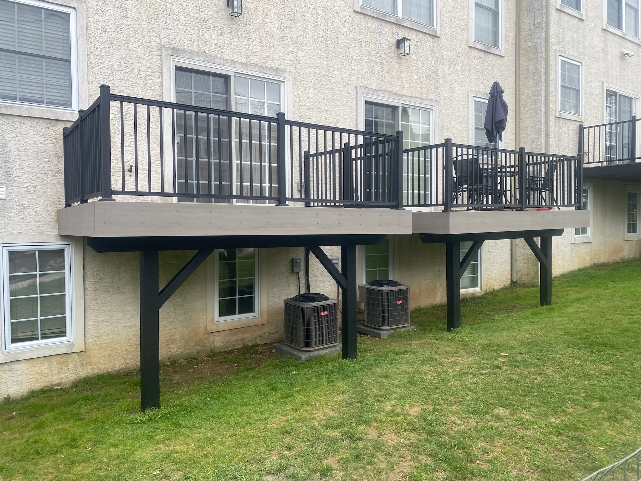 Backyard view of a residential building featuring a balcony with black railings, surrounded by green grass and two HVAC units below.