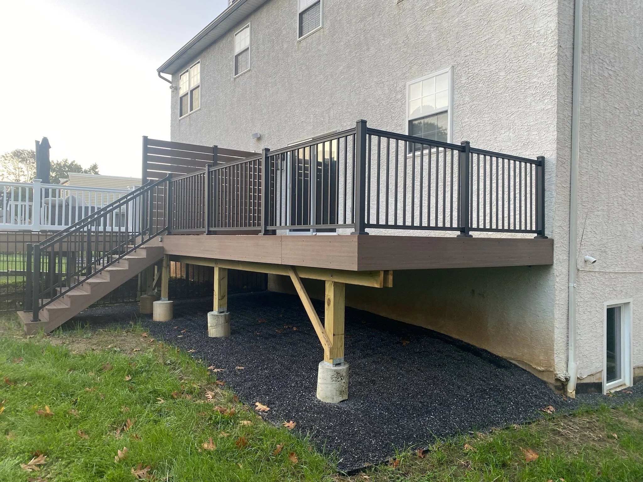 Back of a house with a new wooden deck raised on concrete blocks, with black metal railing, stairs leading up to it, and a sliding glass door.