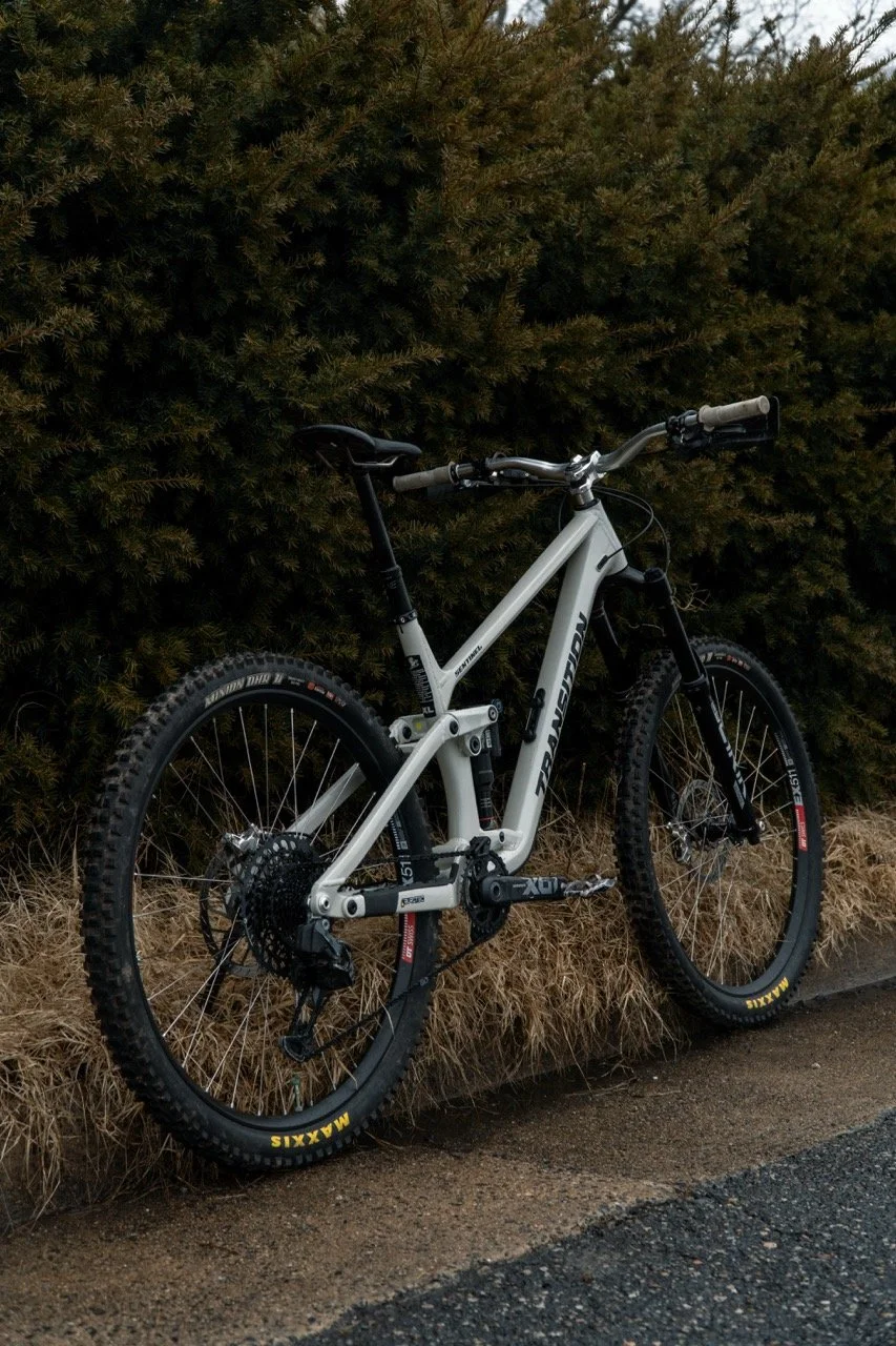 A white Transition Sentinel mountain bike with black tires leaning against a bush with brown grass on the ground.
