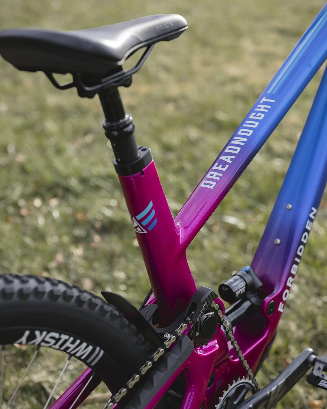 Close-up of a colorful mountain bike with a pink and blue frame, featuring the message 'Dreadnought' on the top tube. The bike has a black seat, chain, and gears, with a background of grass.