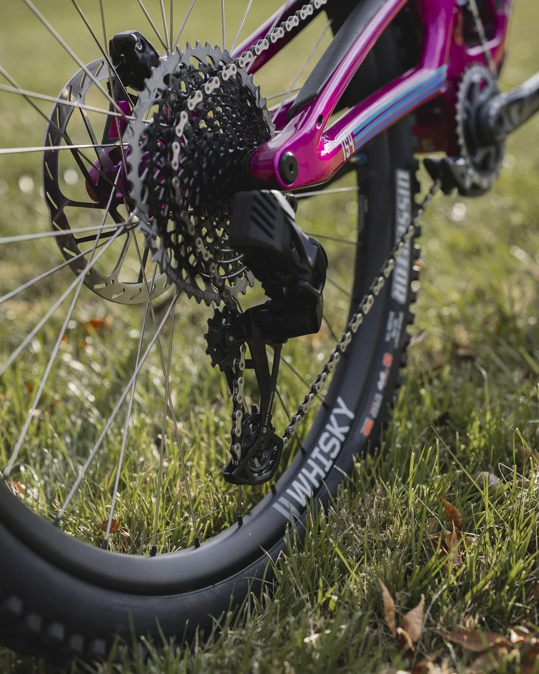 Close-up of a pink mountain bike's rear gear and chain on grass, showcasing the derailleur, cassette, and wheel.