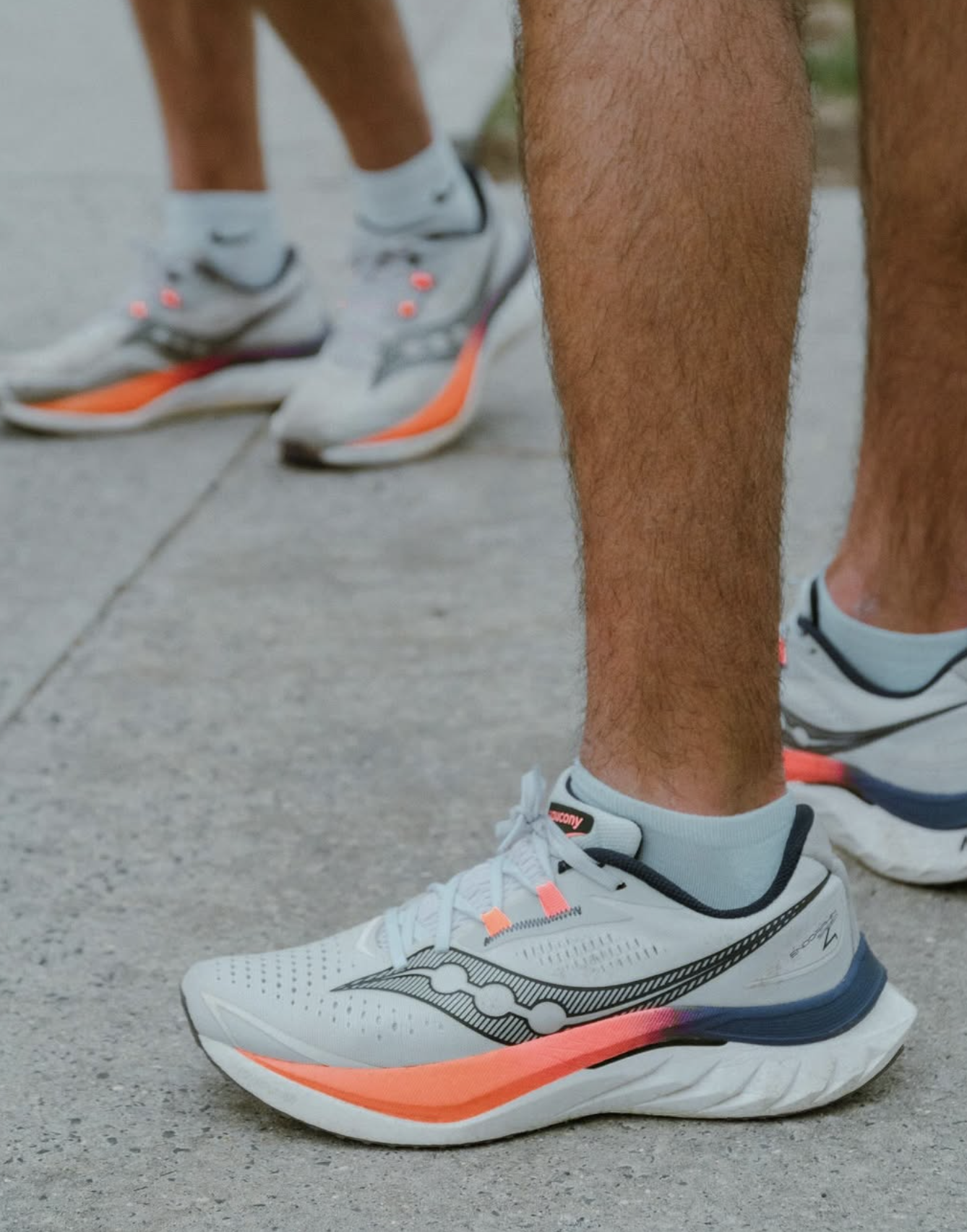 Close-up of a person wearing white and black Saucony running shoes with orange accents, standing on a concrete surface, with another person in similar shoes visible in the background.