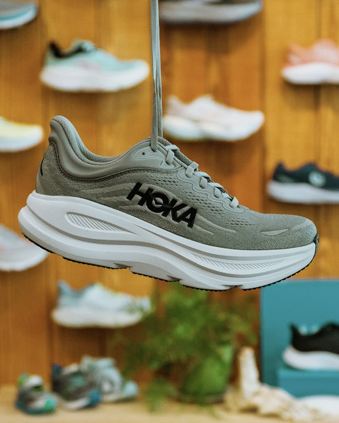 A HOKA Bondi running shoe suspended by a gray shoelace in a shoe store, with other sneakers displayed on wooden shelves in the background.