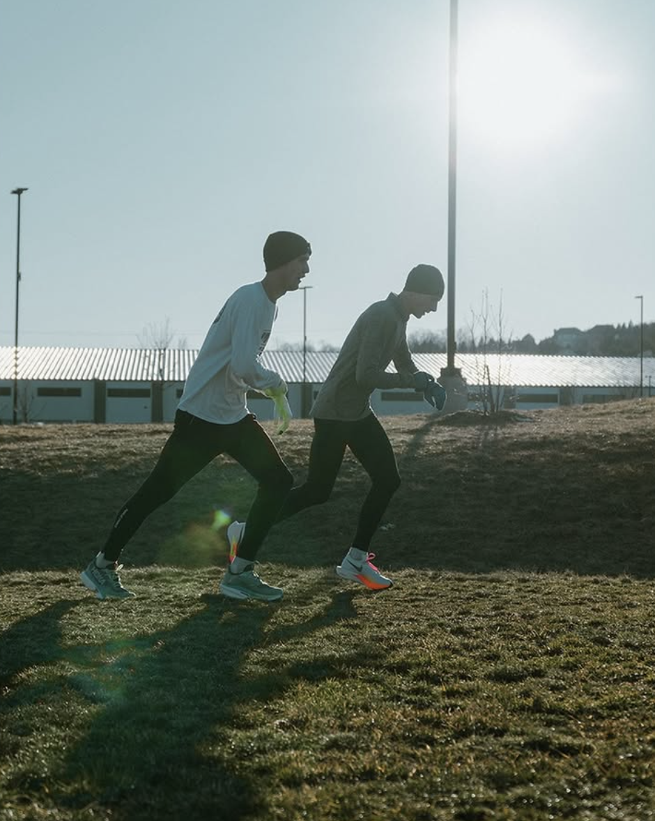 Two young men running outdoors during daylight with the sun shining, on a grassy hill with a fence and buildings in the background.