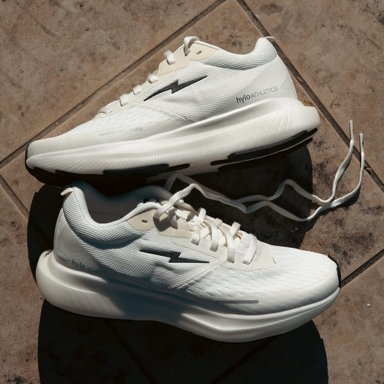 Pair of white HYLO running shoes with black and gray accents on a tiled floor.