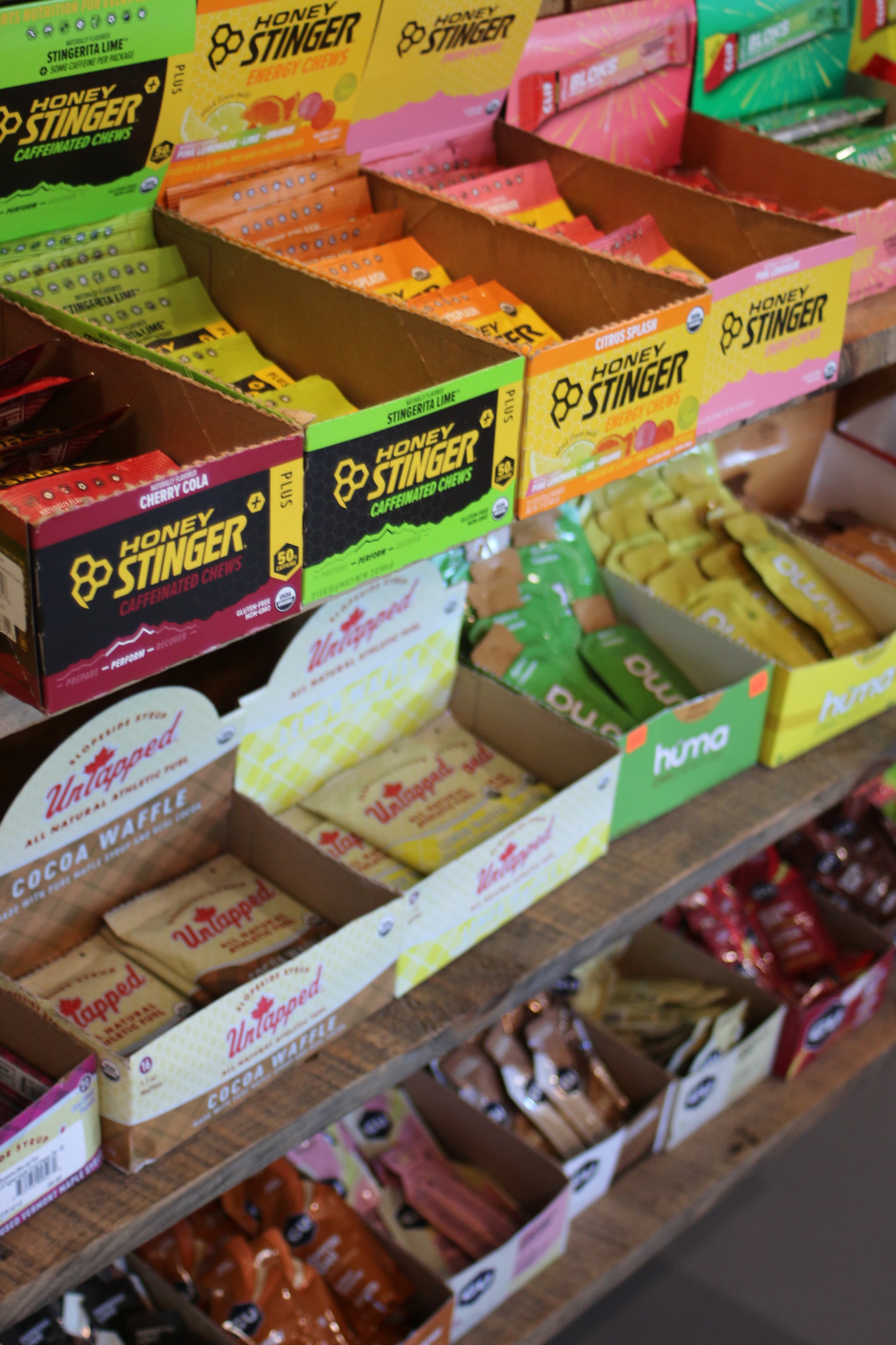 Shelf display of nutrition boxes, including Honey Stinger chews in various flavors, Untapped cocoa waffles, and other ride fuel products.