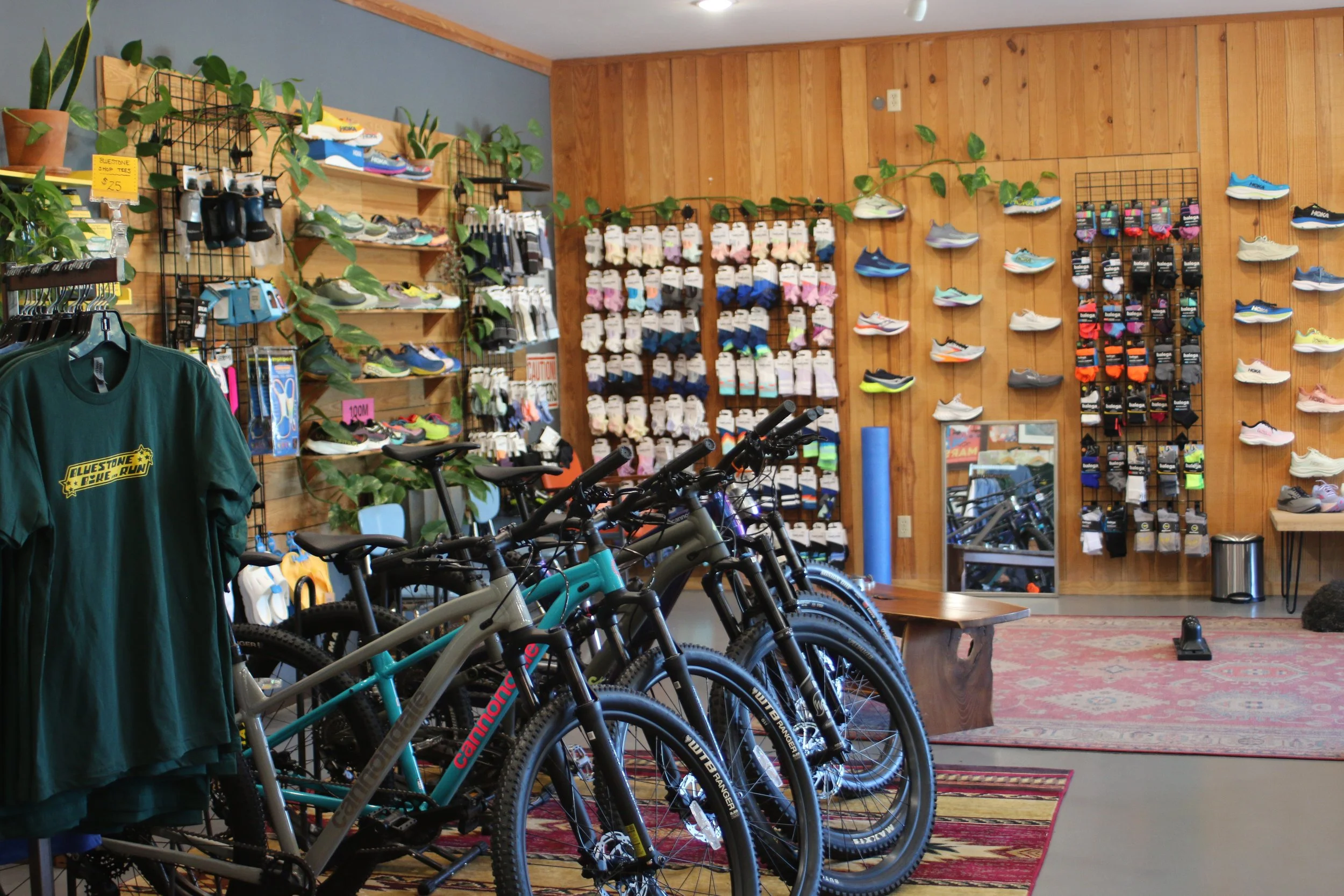 Indoor store with bikes, cycling shoes, and accessories on display. Several bicycles are lined up in the foreground, and shoes and gear are on the wall and shelving.