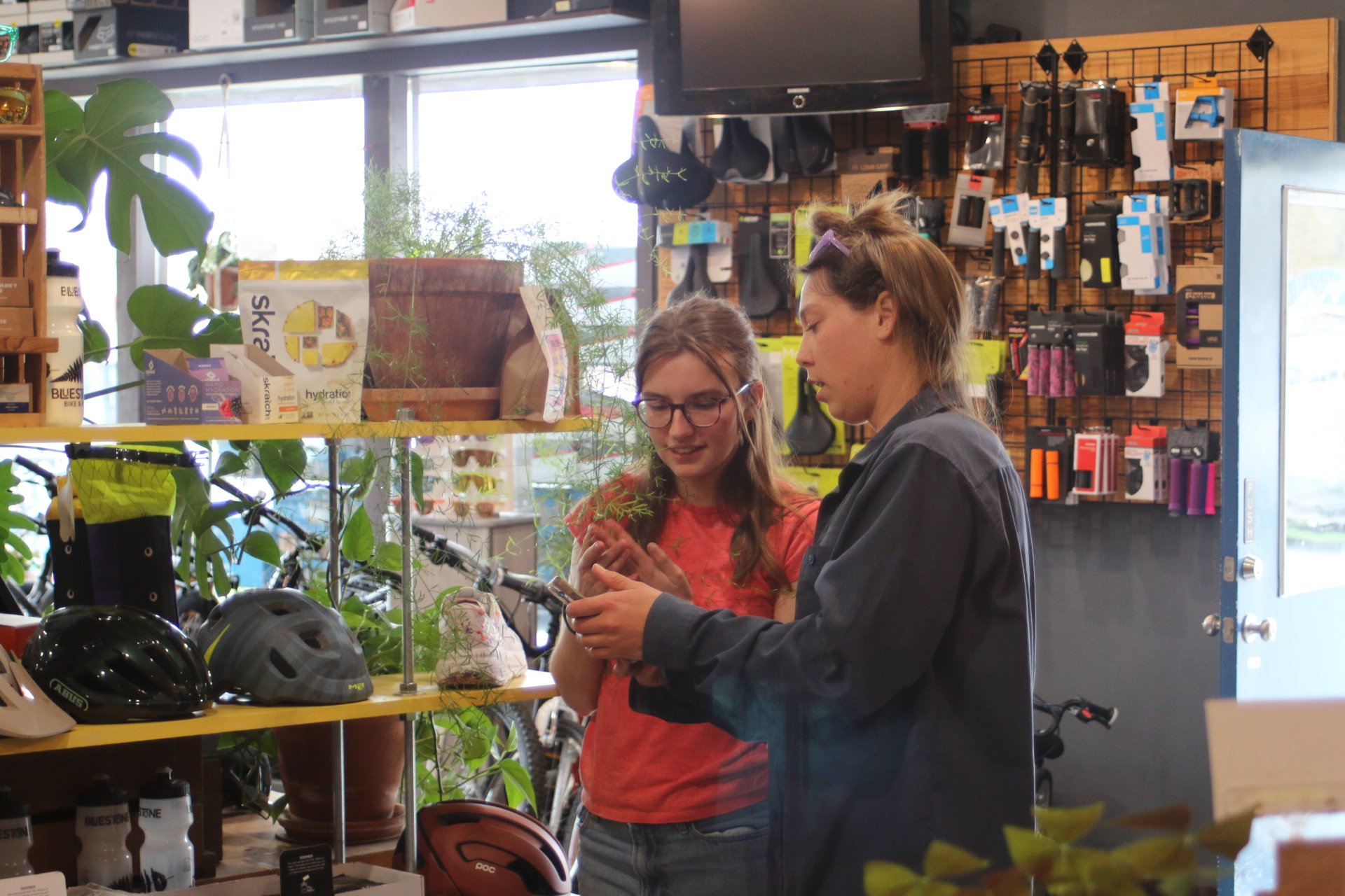 Two women are shopping for bike helmets inside a store. They are looking at a helmet together, surrounded by various helmets and cycling accessories on shelves. The store has a wooden shelving unit with bike parts and a wall with various bike accessories.