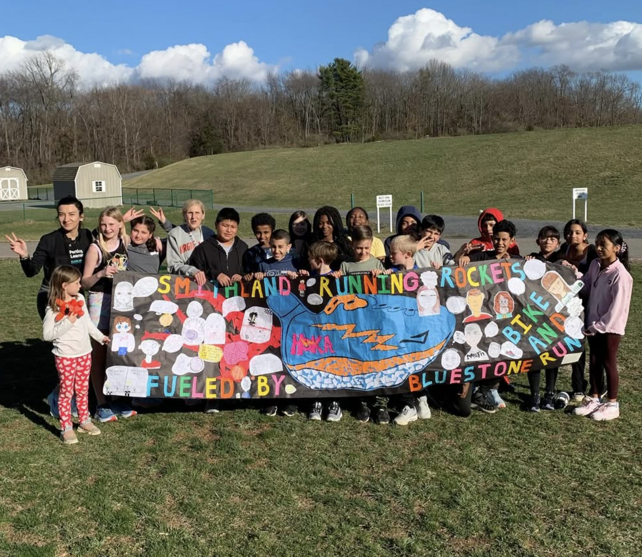Group of children and adults holding a colorful banner outdoors on a grassy field with clear skies and trees in the background.