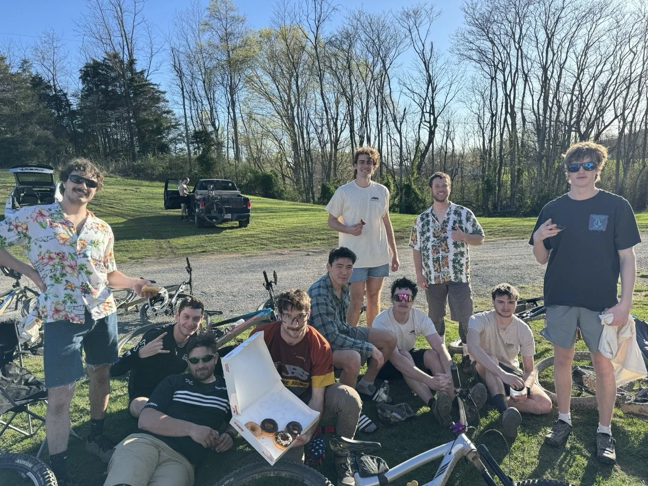 Group of young men outdoors on a sunny day, some sitting on the grass and others standing, with bicycles nearby and a couple of cars in the background, enjoying a casual gathering.