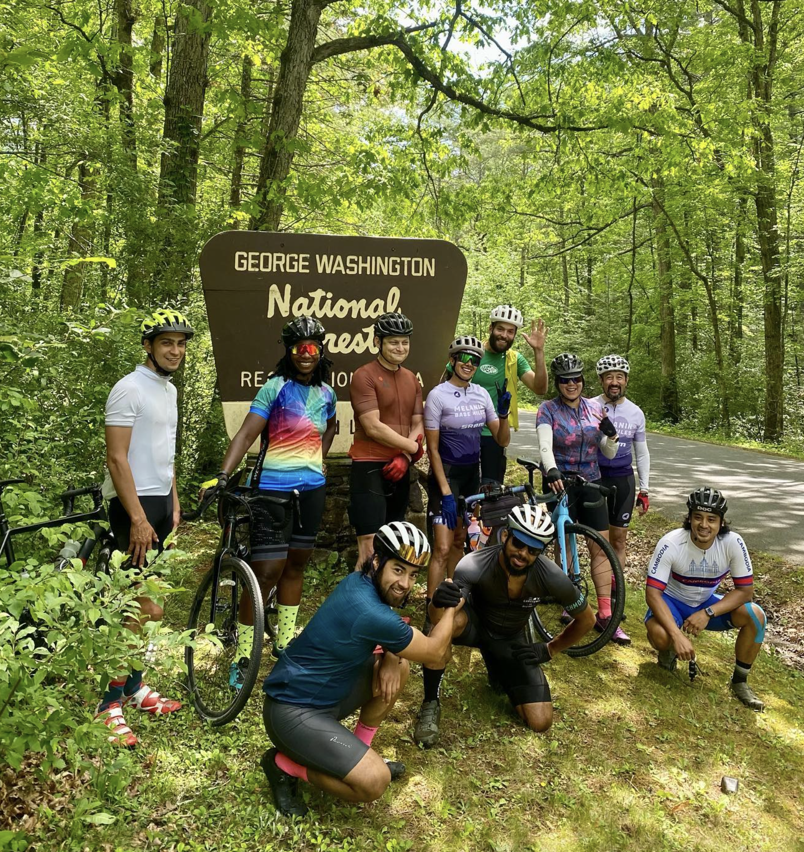Group of cyclists posing next to a sign that reads 'George Washington National Forest,' in a lush green forested area, some holding bikes, wearing helmets and cycling gear, smiling and making gestures.