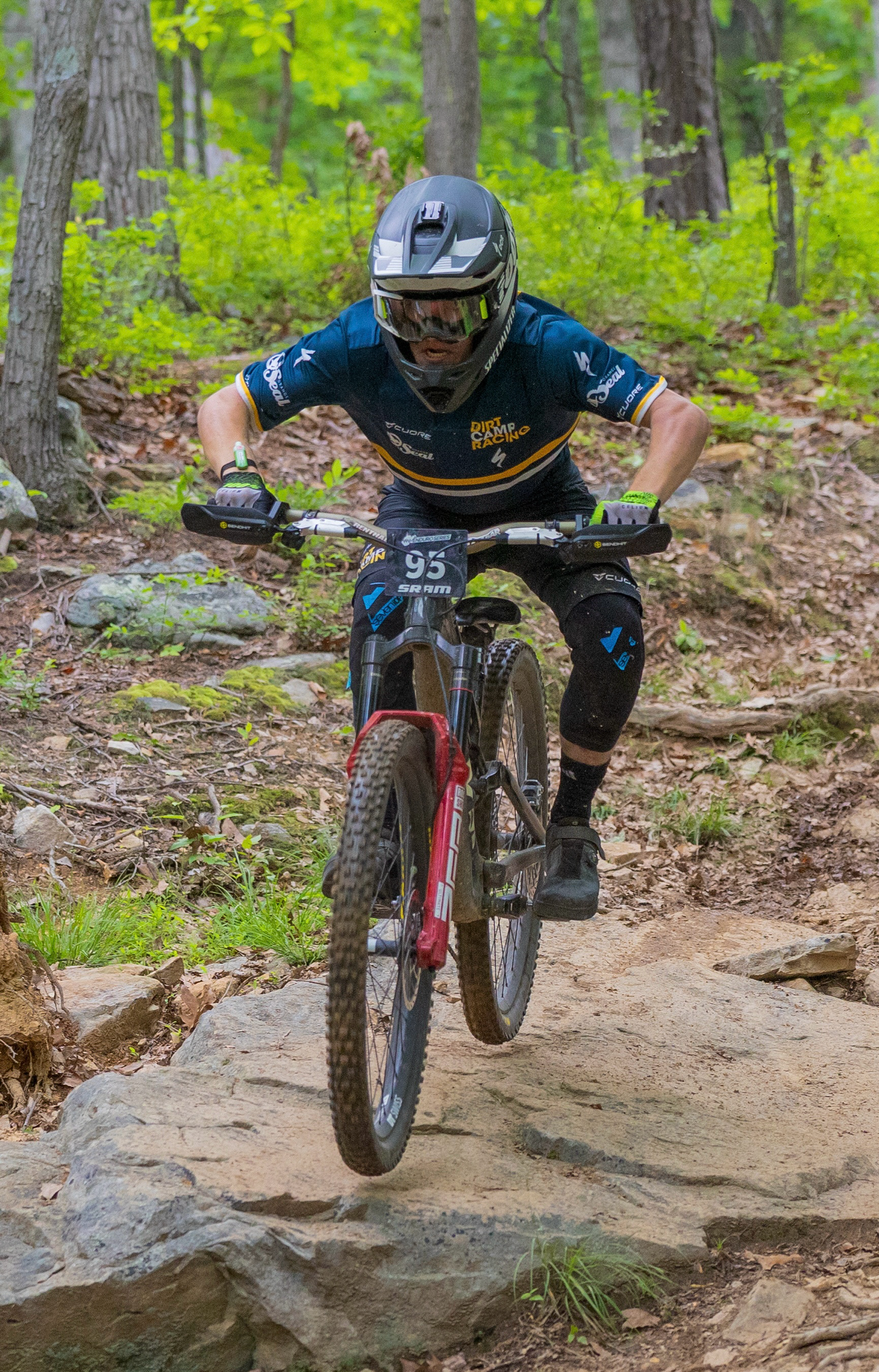 Mountain biker wearing a helmet and racing gear riding over a large rock on a dirt trail in a wooded area.