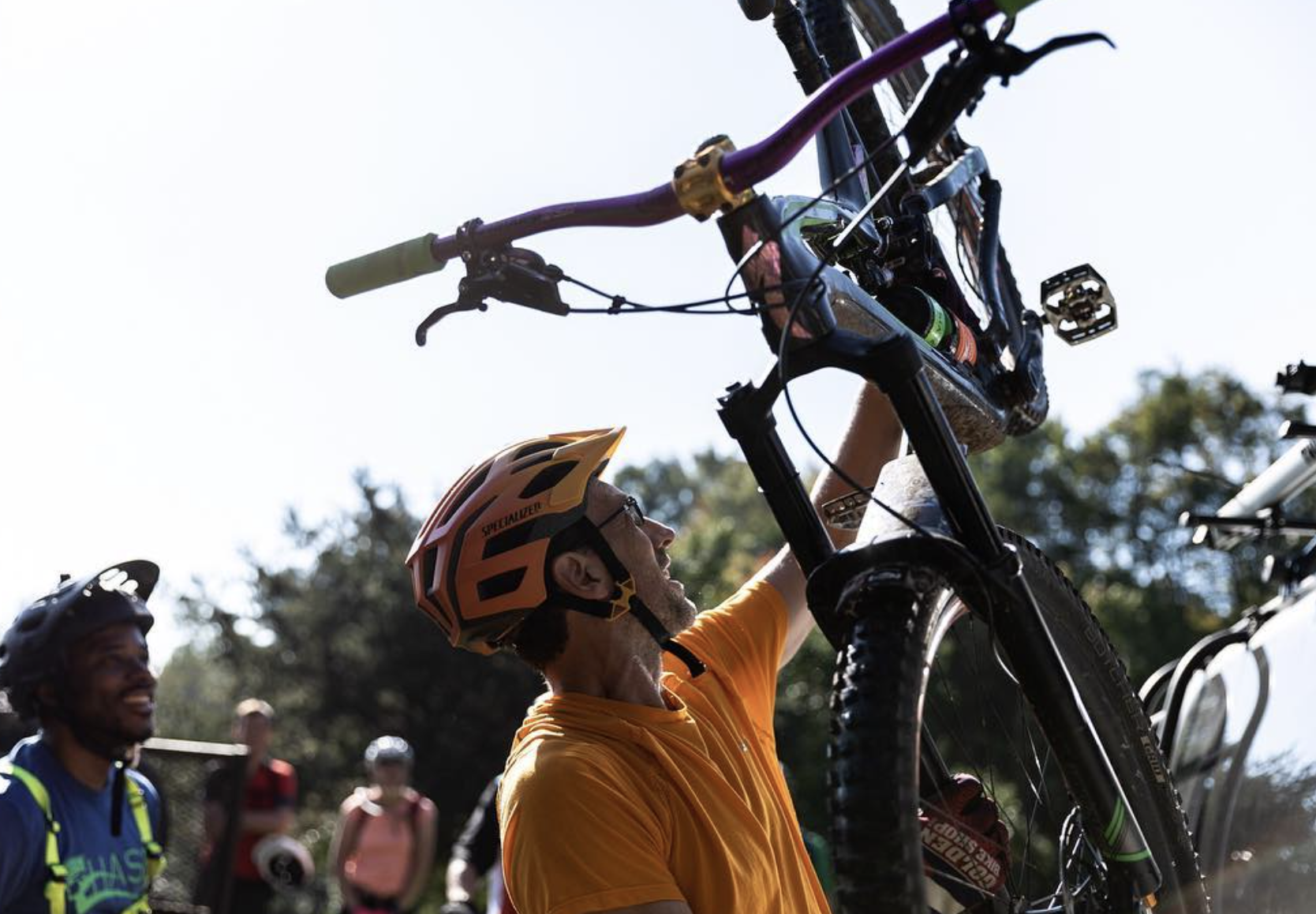 Man wearing an orange shirt and a bike helmet holding a mountain bike above his head, with other bikers in the background.