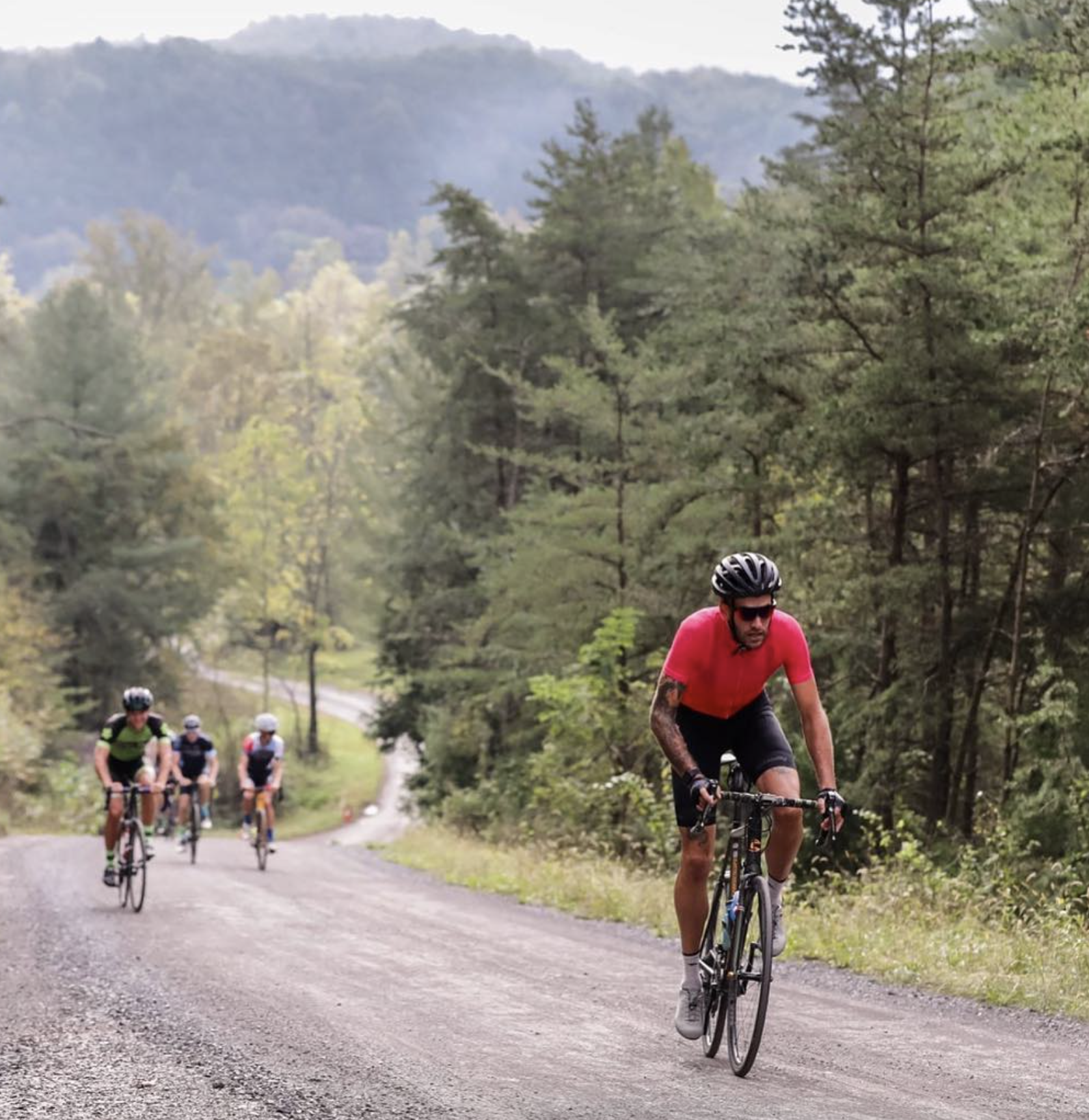 Four cyclists riding on a dirt trail surrounded by green trees and distant mountains in the background.